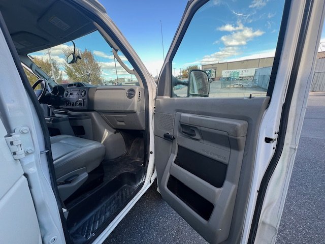 Open driver-side door of a white utility truck showing the gray interior, dashboard, and part of the front seat, with a clear sky and industrial buildings in the background.