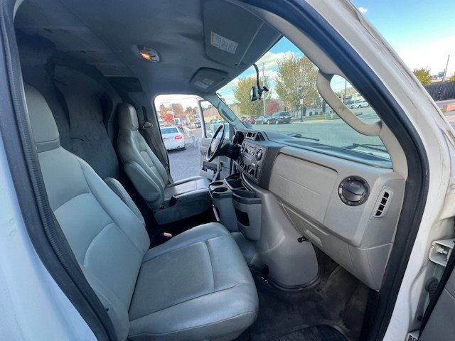 Interior of a commercial van showing the driver and passenger seats, dashboard, and steering wheel, with a view of a parking lot outside through the windshield.