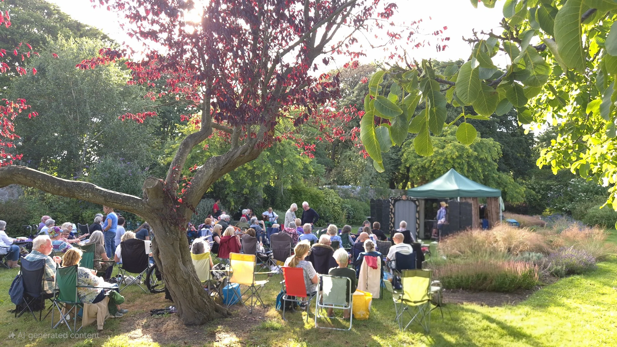 A large outdoor gathering of people seated on chairs in a garden with trees, some standing and talking, with a small stage or shelter in the background and sunlight filtering through the trees.