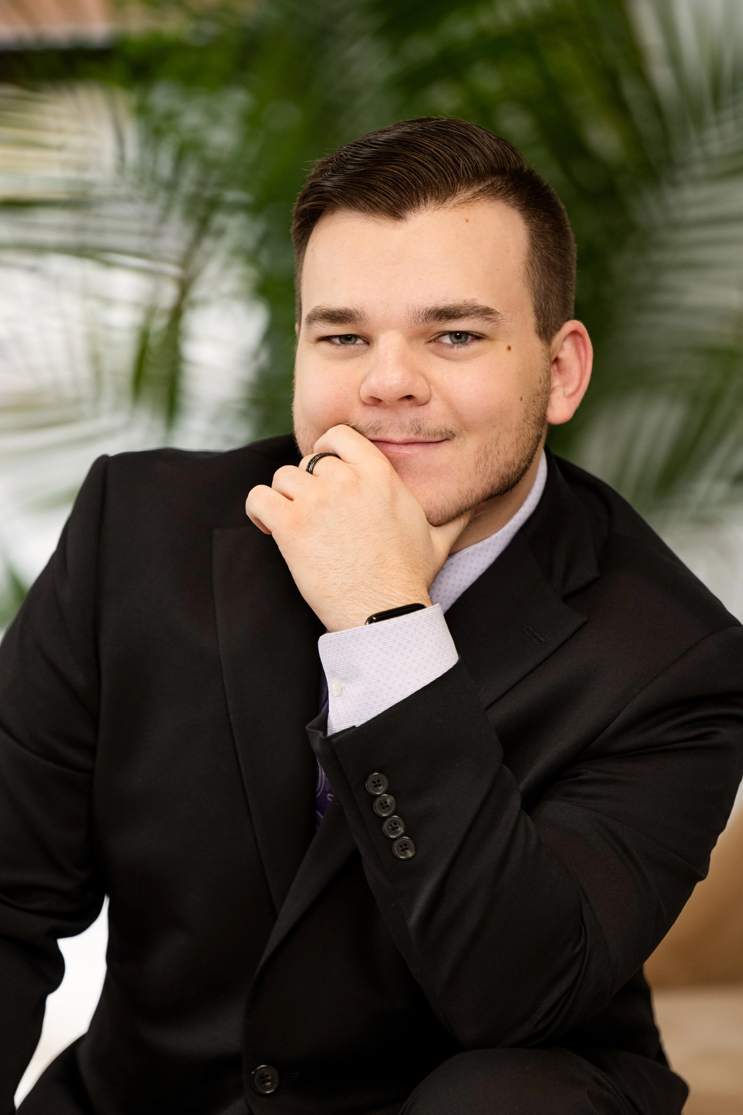A young man in a black suit with a light blue shirt, sitting thoughtfully with his hand near his face, indoors with green plants in the background.