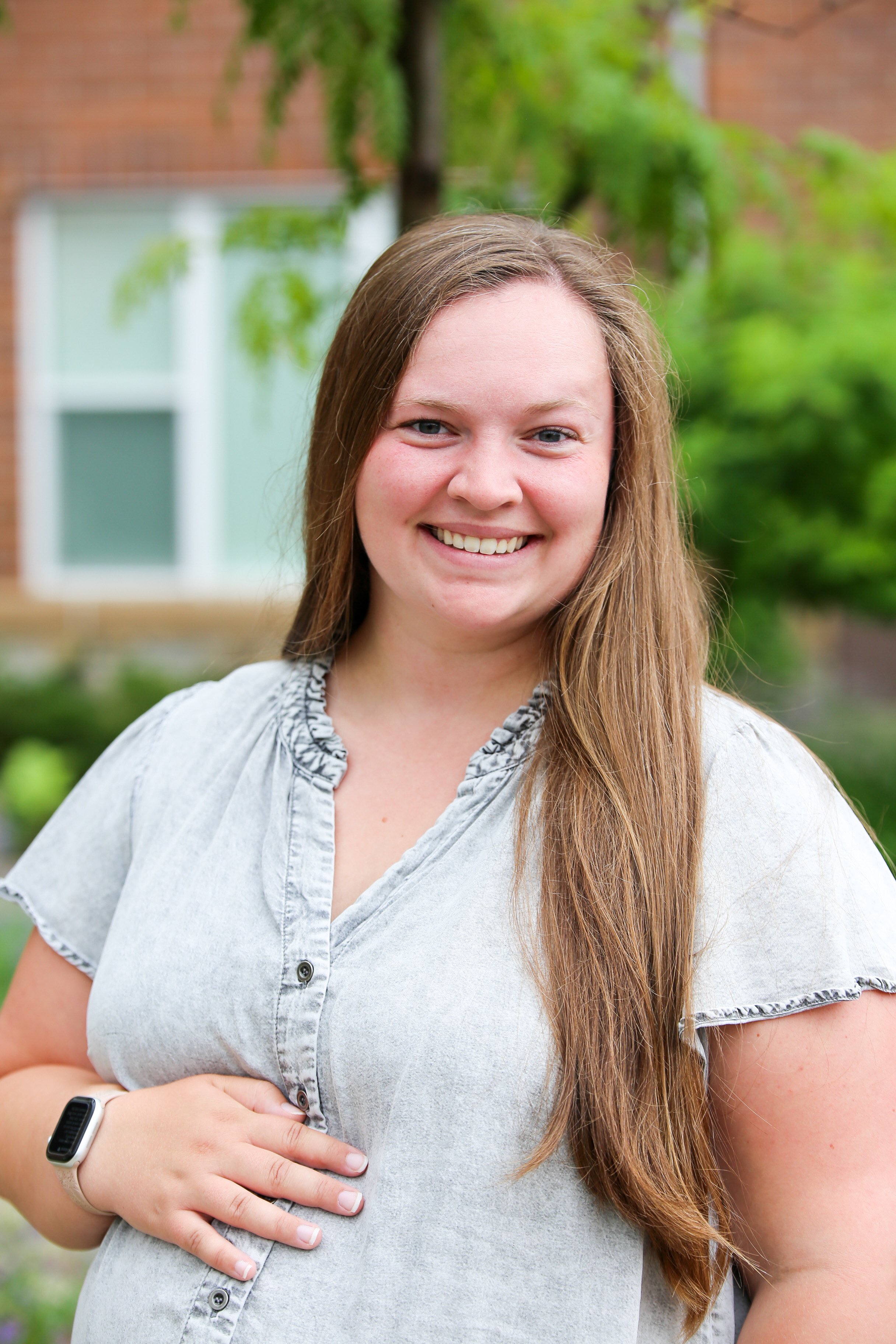 A pregnant woman with long brown hair smiling outdoors on a sunny day, standing on a dirt path surrounded by greenery and trees.