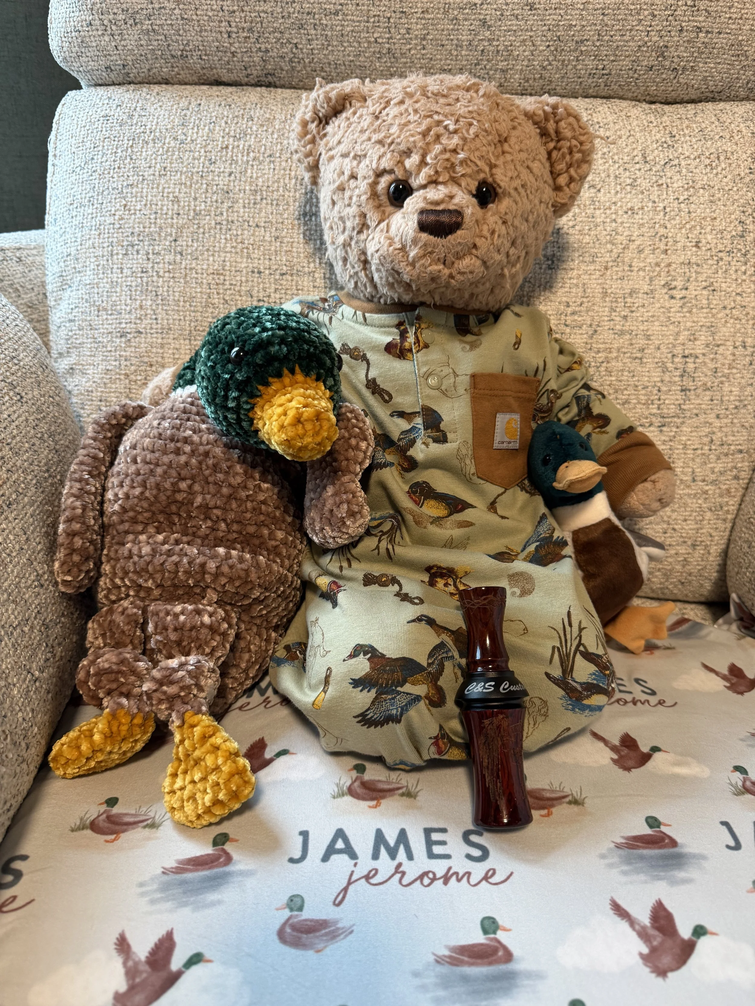 Stuffed teddy bear with a plush duck toy seated on a blanket with duck and goose patterns, next to a plush duck, on a couch.