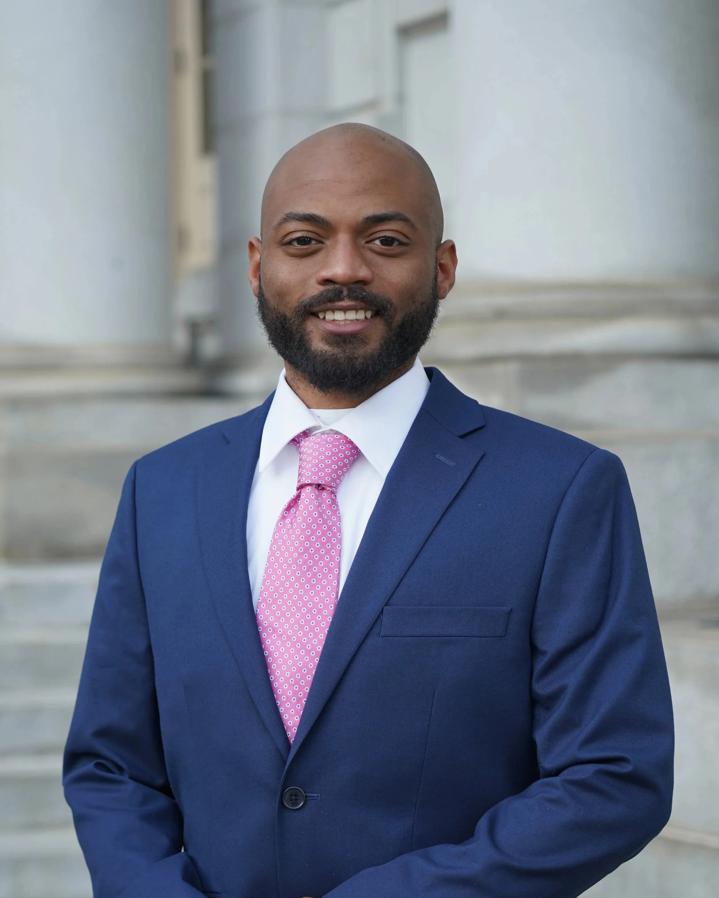 A man wearing a dark blue suit, white shirt, and pink tie, standing outdoors in front of a stone building with steps.