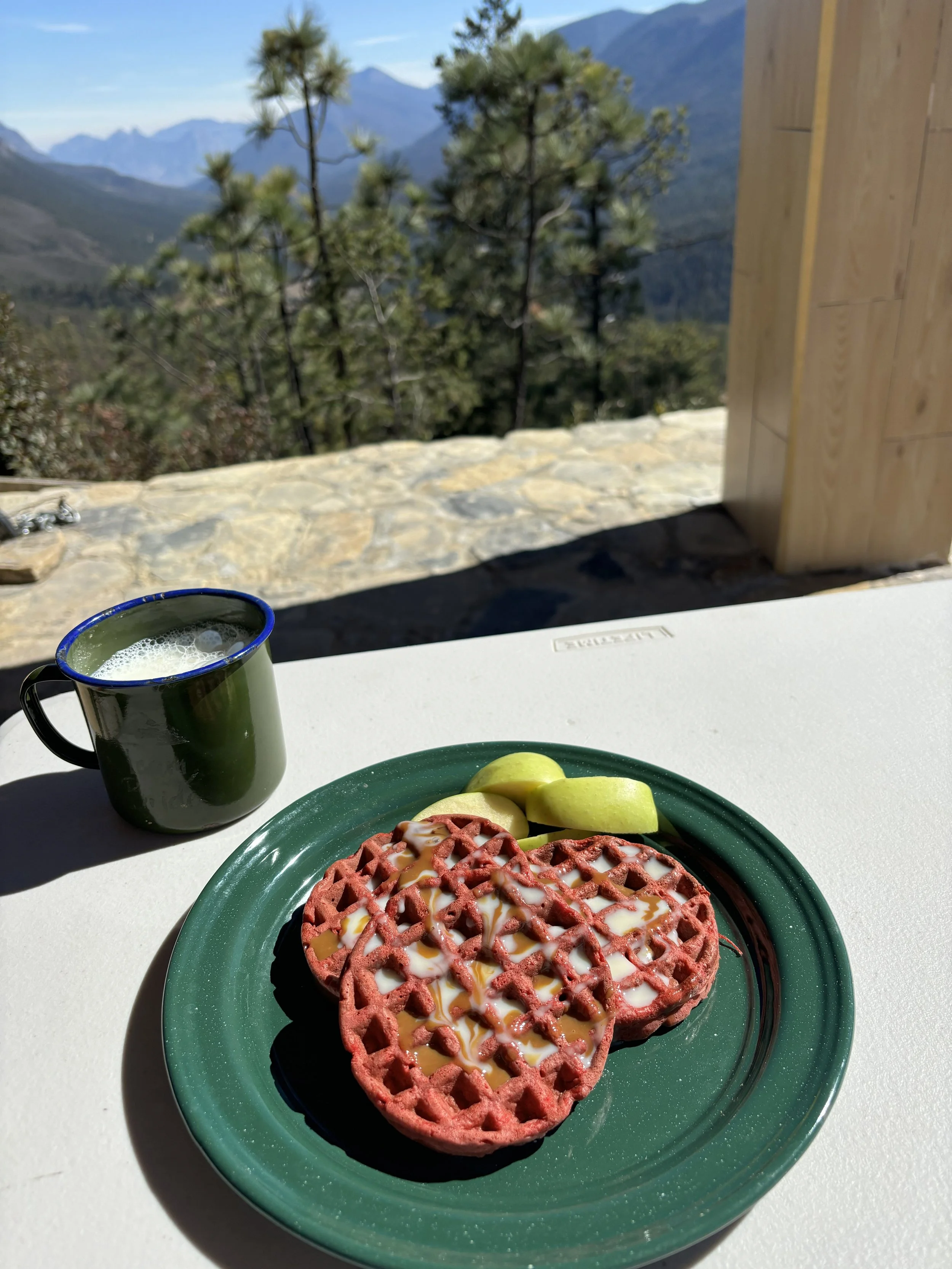 A plate of red waffles with white icing and syrup, served with apple slices, a green mug of coffee or tea, on a white table, with a mountain landscape view in the background.