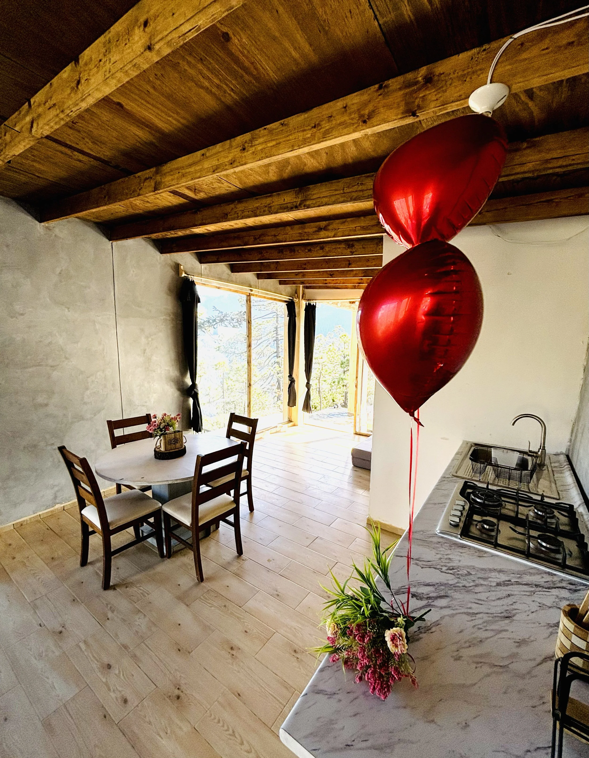A kitchen with wooden ceiling beams, a round dining table with four chairs, large windows with black curtains, and two red heart-shaped balloons tied to a vase with flowers on a marble countertop.