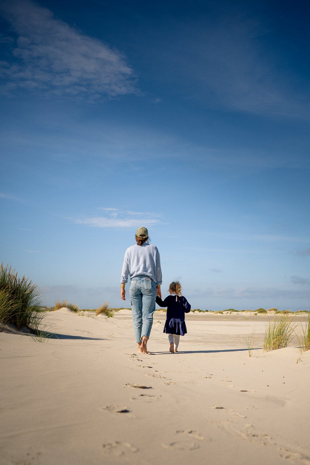 Eine erwachsene Frau und ein kleines Mädchen gehen barfuß am Strand entlang, umgeben von Sand und Dünen, bei strahlend blauem Himmel.