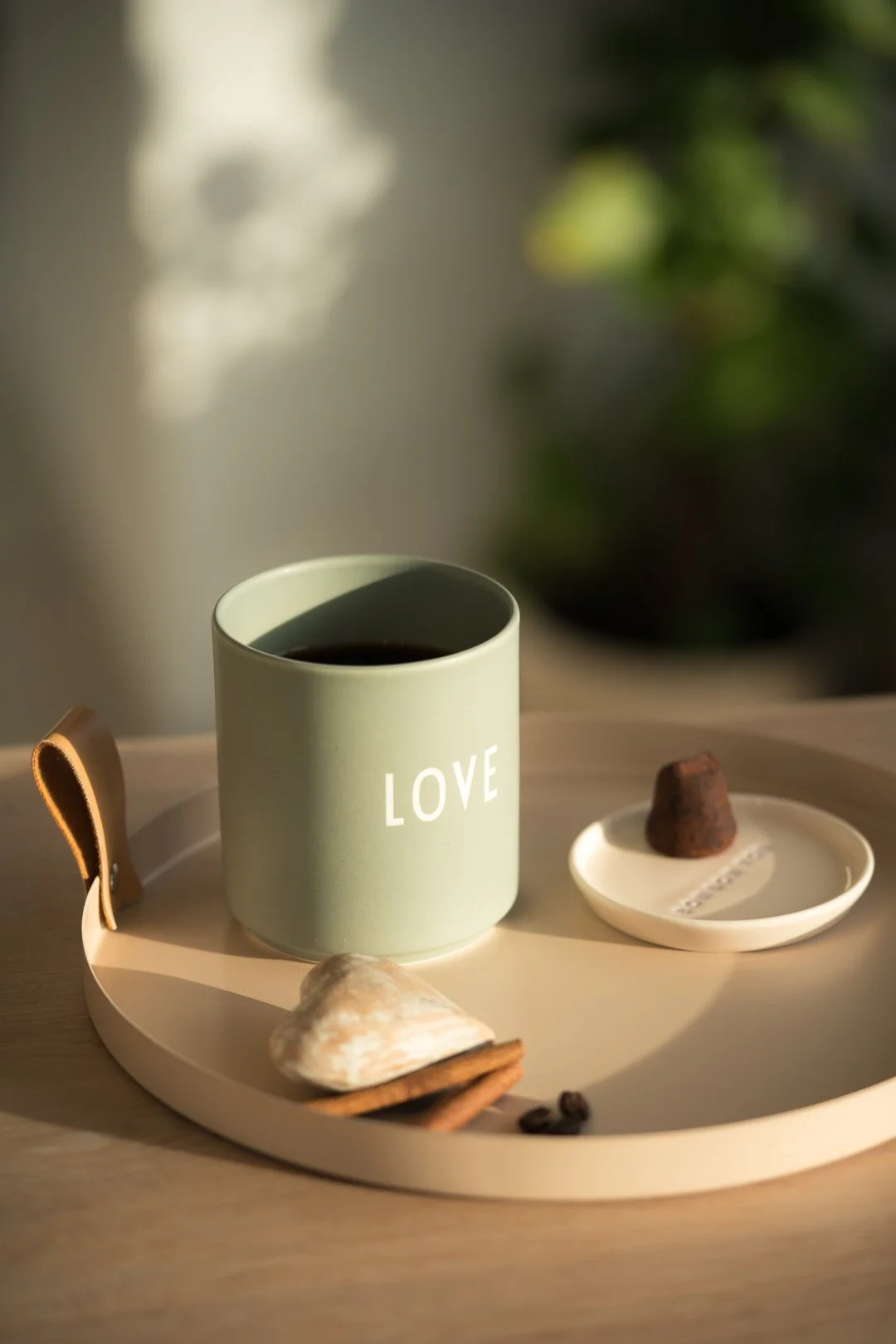 Eine Tasse mit dem Wort "LOVE" darauf, auf einem runden beige Tablett, daneben eine kleine weiße Schale mit einer Praline, ein Herz aus Muscheln und Zimtstangen, im Hintergrund grüne Pflanzen und Sonnenlicht.