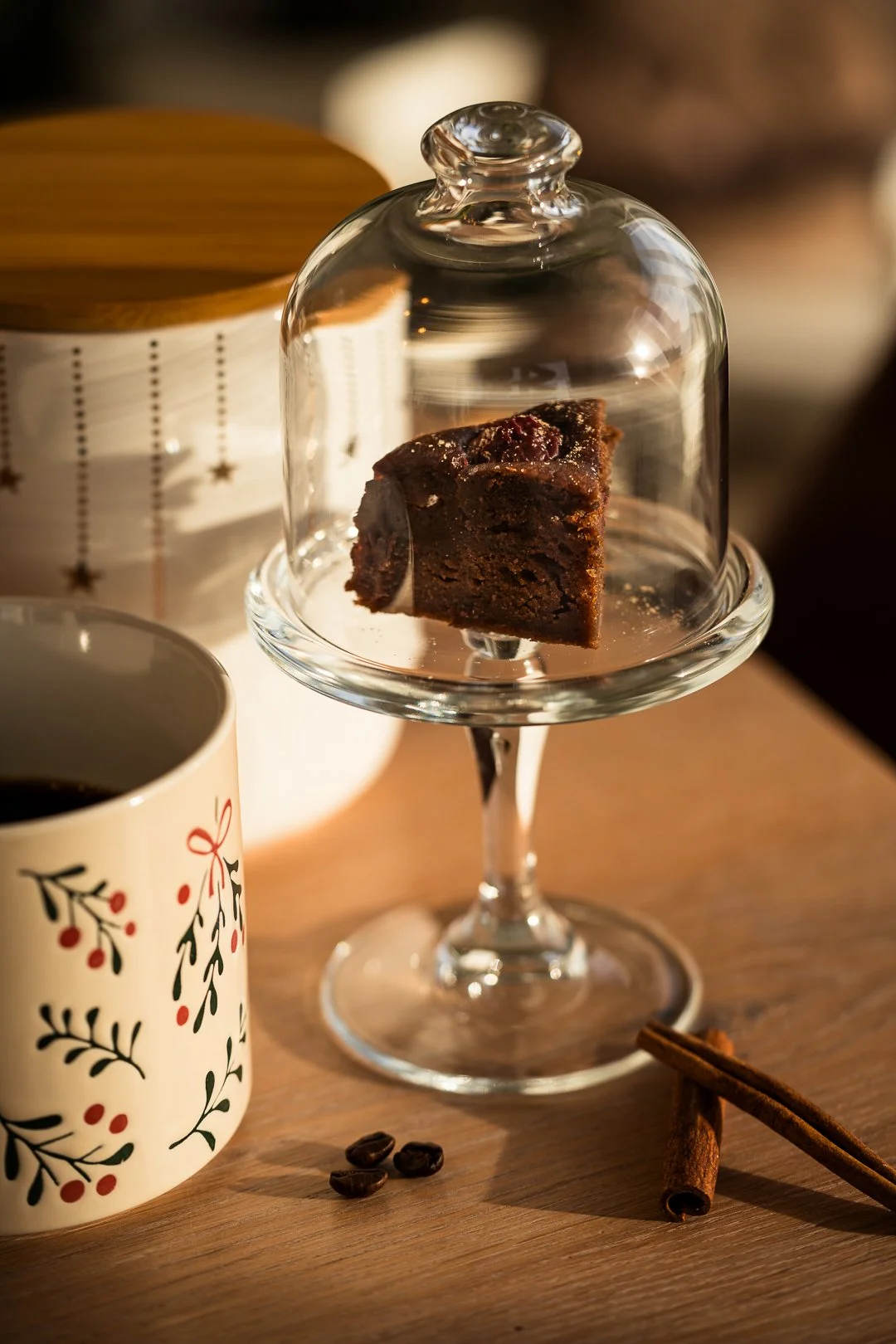 Schwarzer Kuchenkuchen auf einem Glasdessertständer, umgeben von Kaffeetassen, Zimtstangen und Kaffeebohnen, auf einem Holztablett.