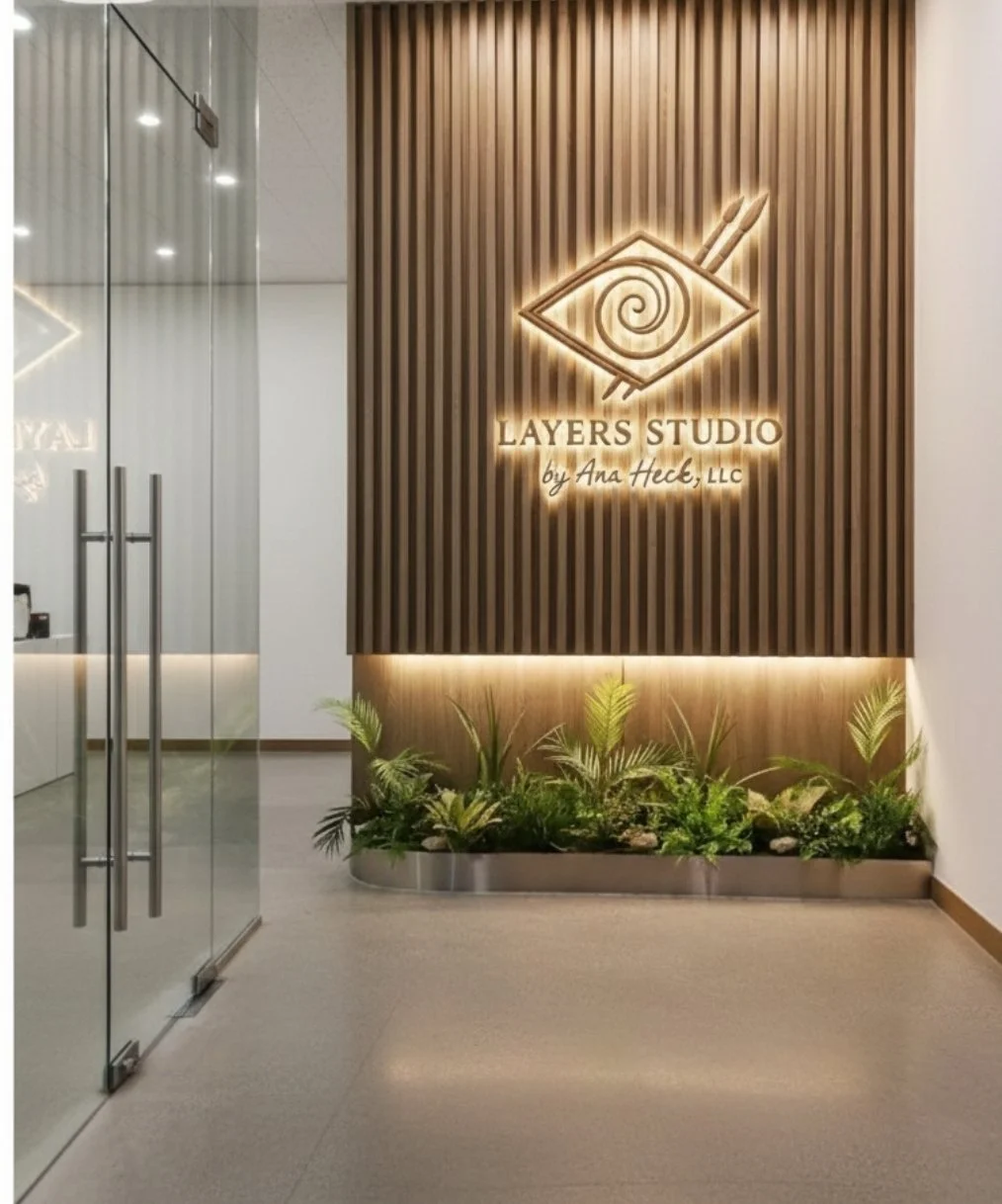 Office lobby with a wooden accent wall featuring a backlit logo for Layers Studio by Ana Heck, LLC, and green plants at the base of the wall.