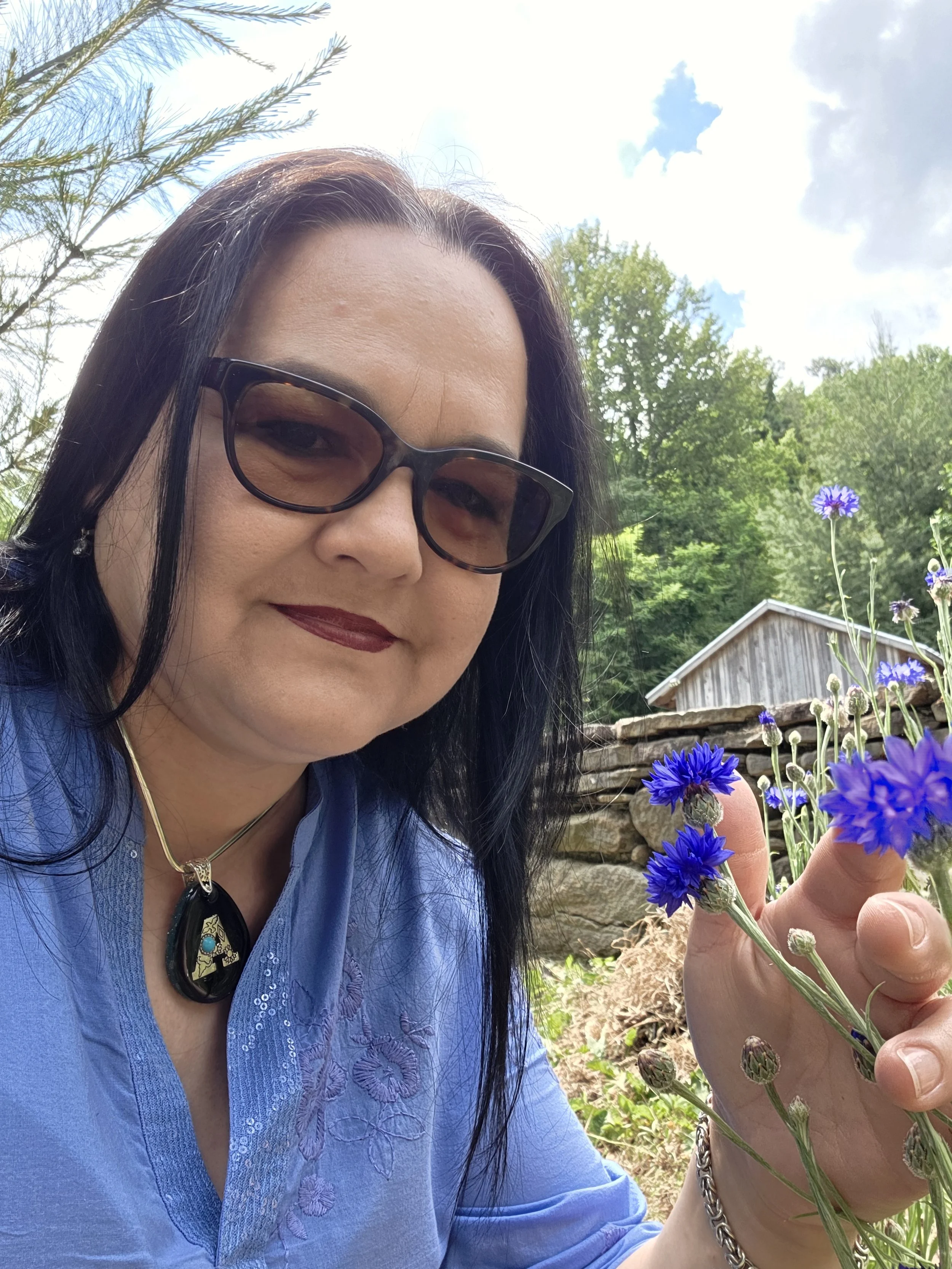 A woman with dark hair, wearing glasses and a blue embroidered shirt, is holding a bunch of purple flowers outdoors with trees and a wood building in the background.