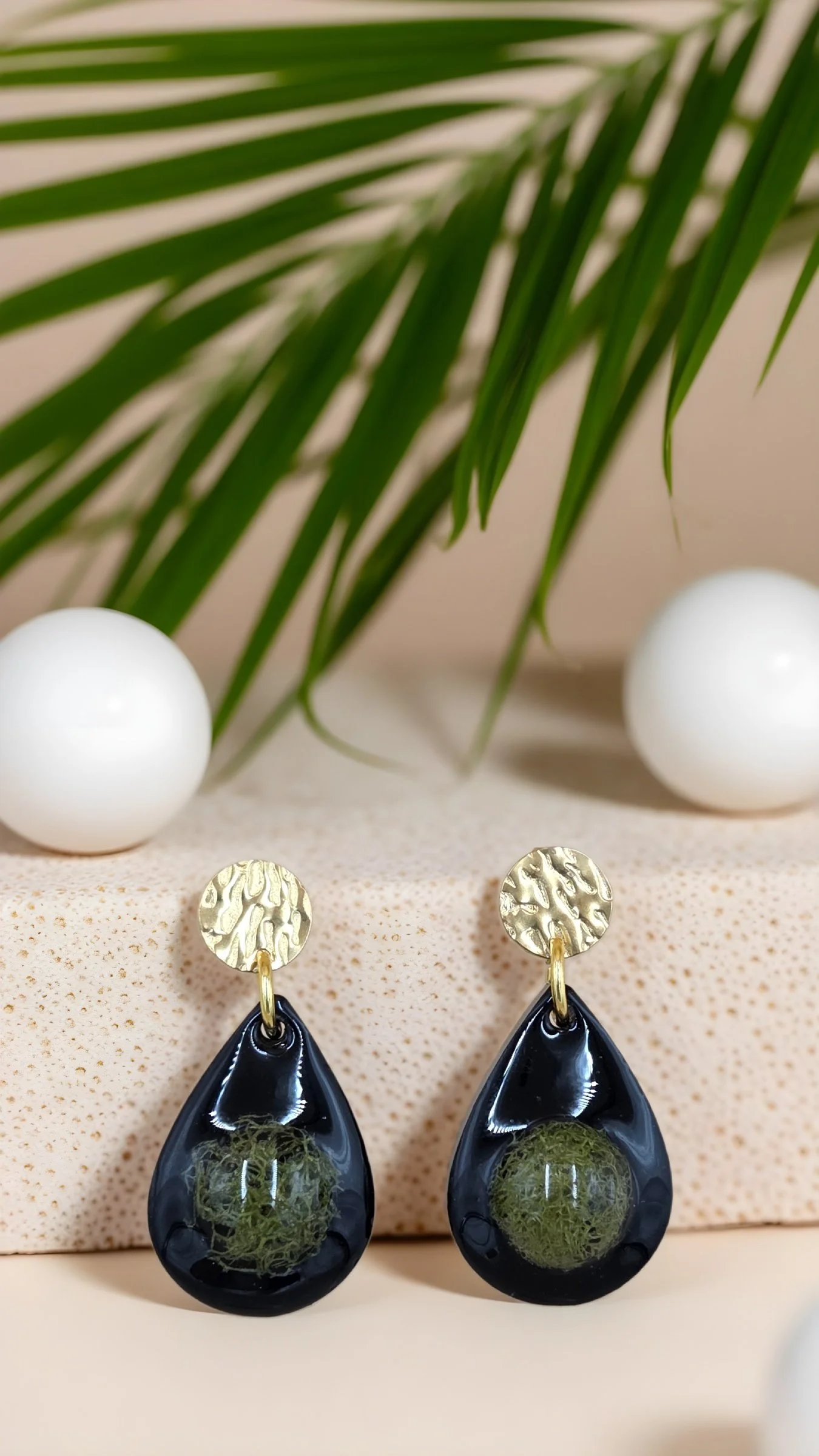 Black teardrop-shaped earrings with green moss inside, hanging from textured gold studs, placed on a white speckled surface with large white and green palm fronds in the background.
