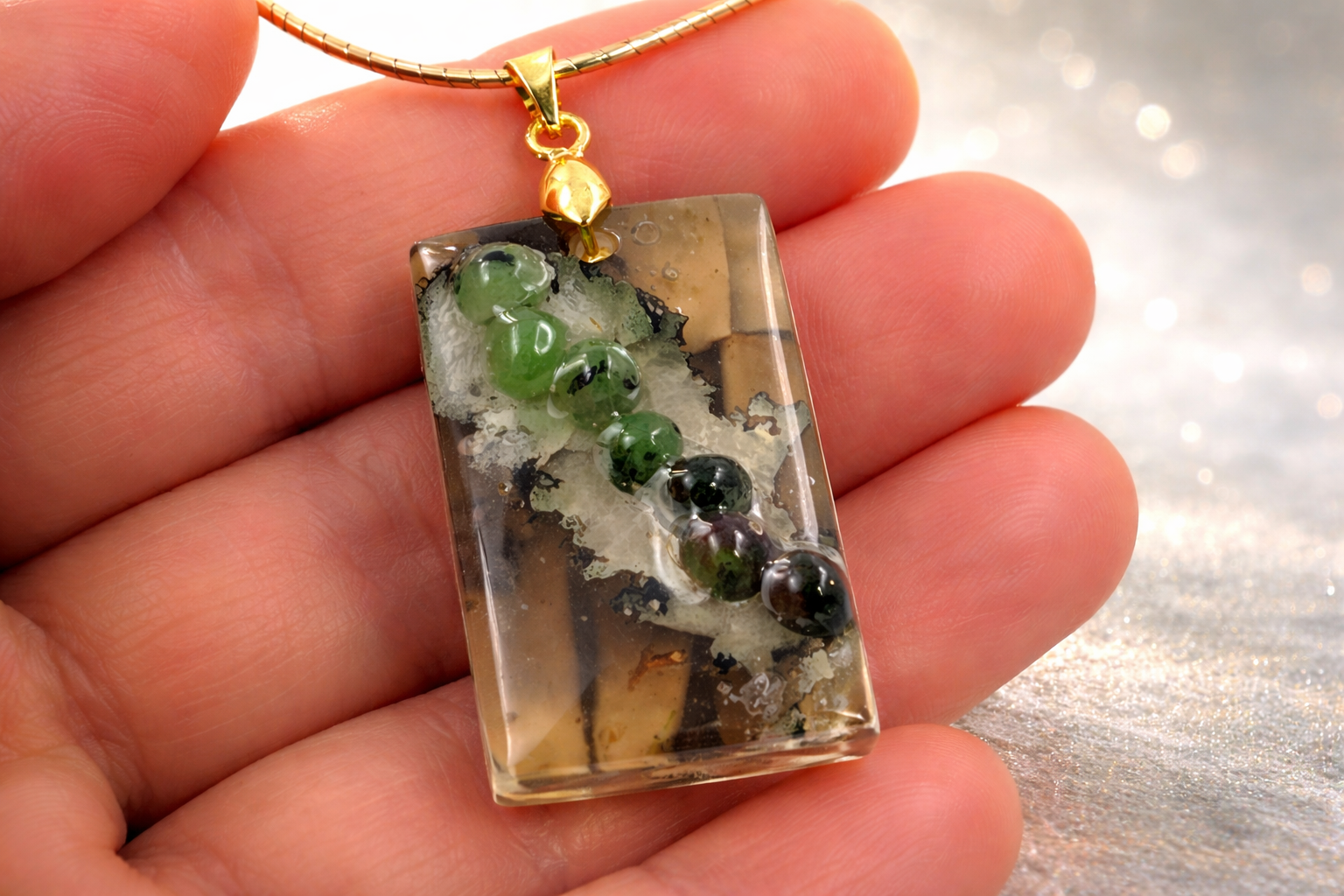 A close-up of a person's hand holding a pendant with green lichen and dark beads embedded in clear resin, attached to a gold chain.