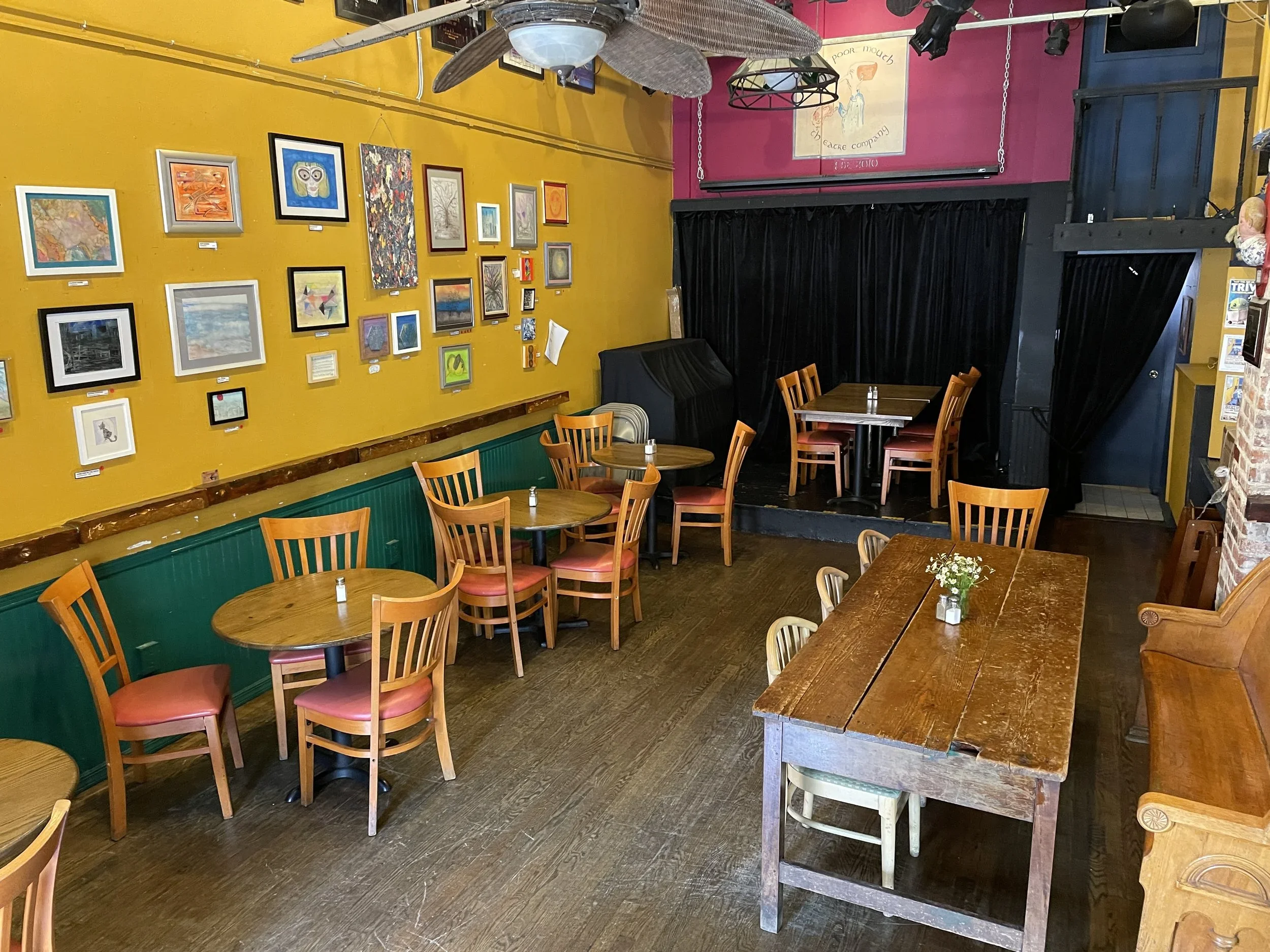Interior of a cozy cafe with colorful walls, featuring an art gallery with various framed artworks on a yellow wall. Wooden tables and chairs are arranged for dining, and a small stage with black curtains is at the back.