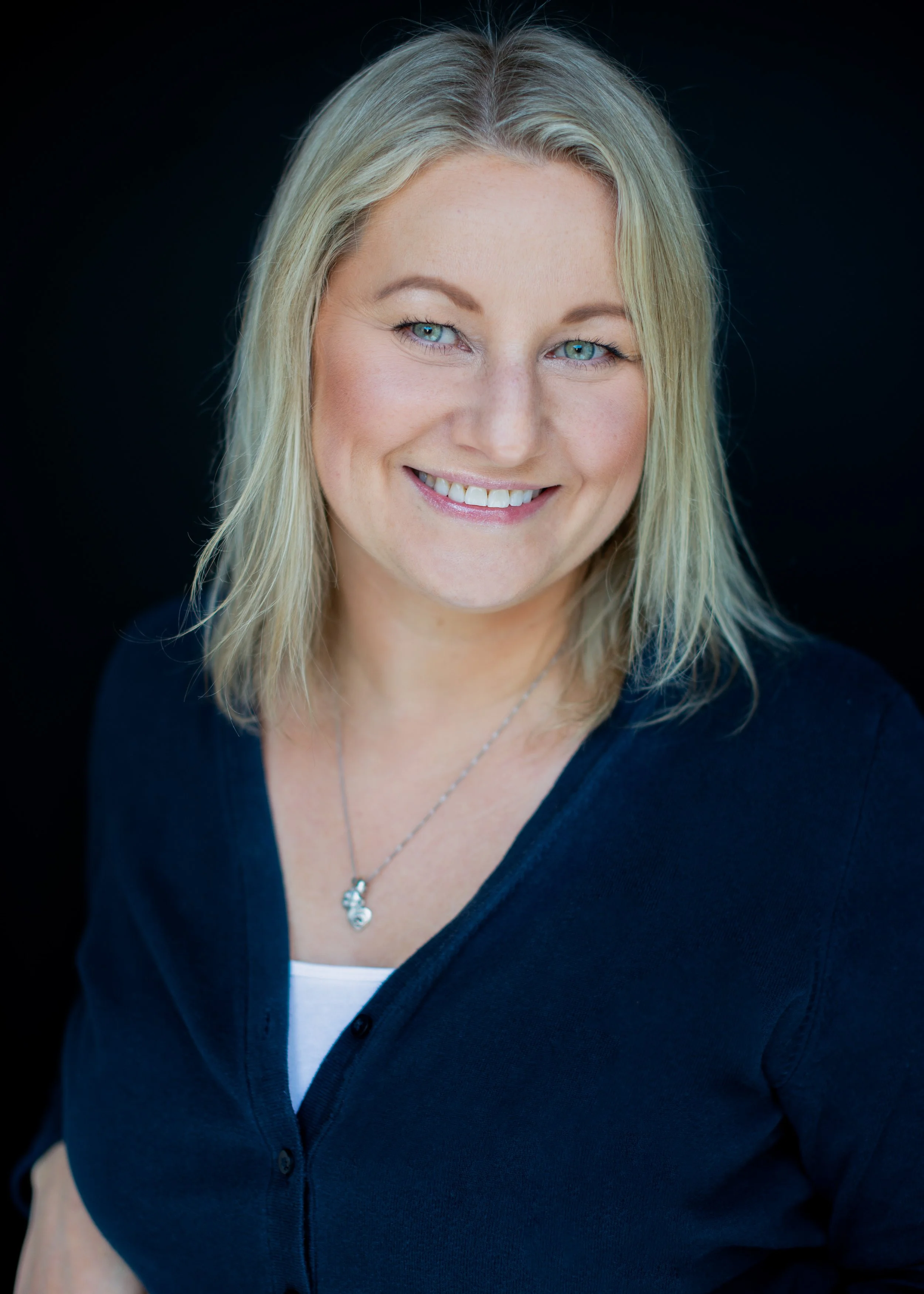 Headshot of a smiling blonde woman with blue eyes wearing a dark blue top and a silver necklace, against a black background.