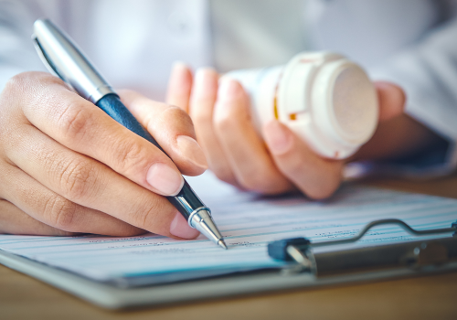 A person writing on a clipboard with a pen, with a prescription bottle nearby.