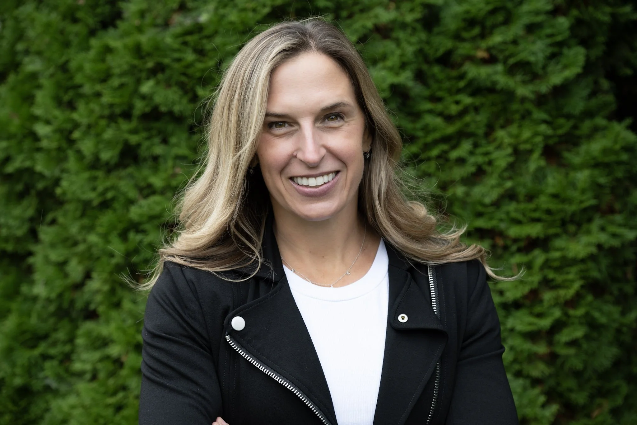 A woman smiling with long, wavy blond hair, wearing a black jacket over a white top, standing outdoors against a backdrop of green foliage.
