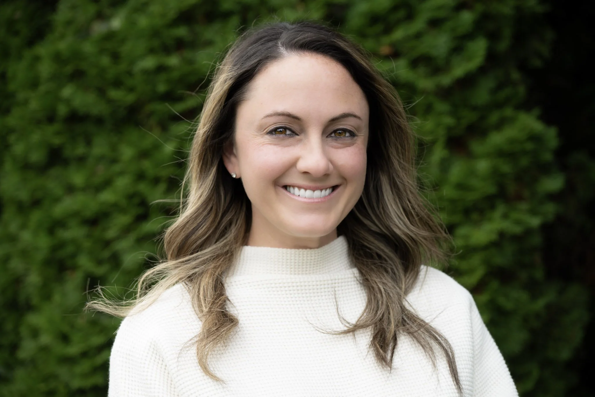 A woman with shoulder-length wavy brown hair smiling outdoors, wearing a white textured sweater, in front of a green leafy background.