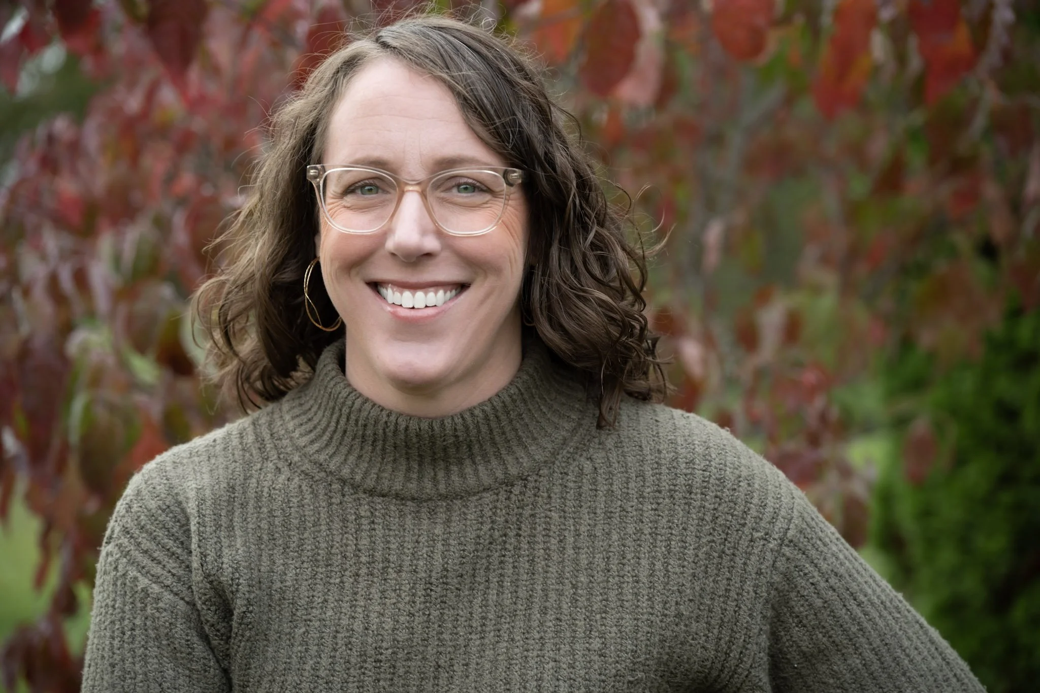 Woman with curly brown hair wearing glasses, hoop earrings, and a green turtleneck sweater, smiling outdoors with red and green foliage in the background.