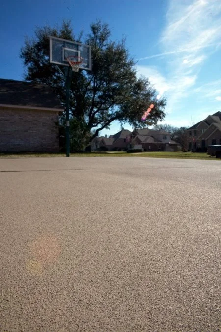 Empty suburban outdoor basketball court with a basketball hoop, trees, and houses in the background under a blue sky.