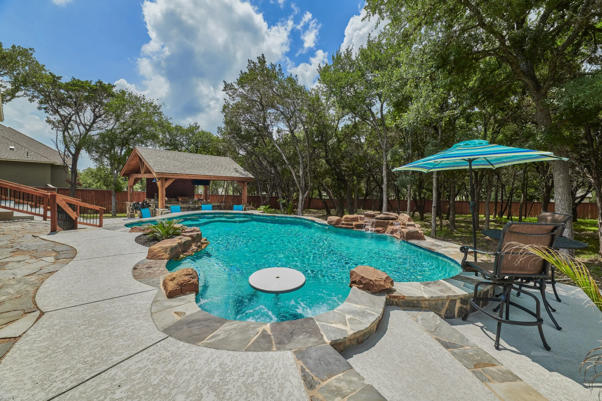 Backyard swimming pool with a stone border, shaded seating area with a blue striped umbrella, and surrounded by trees and a wooden fence.
