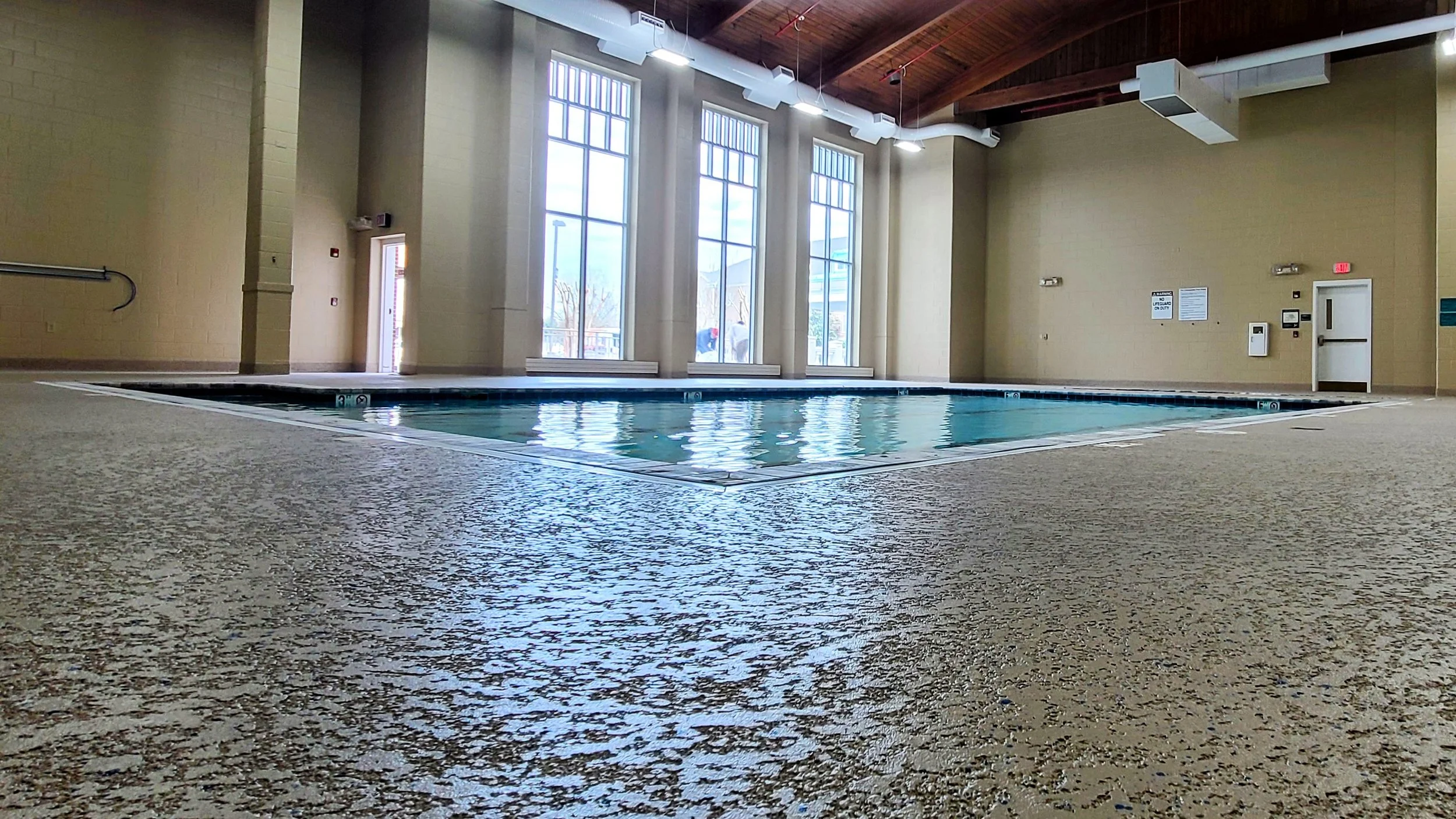Indoor swimming pool with large windows allowing natural light, beige walls, and a textured beige floor.