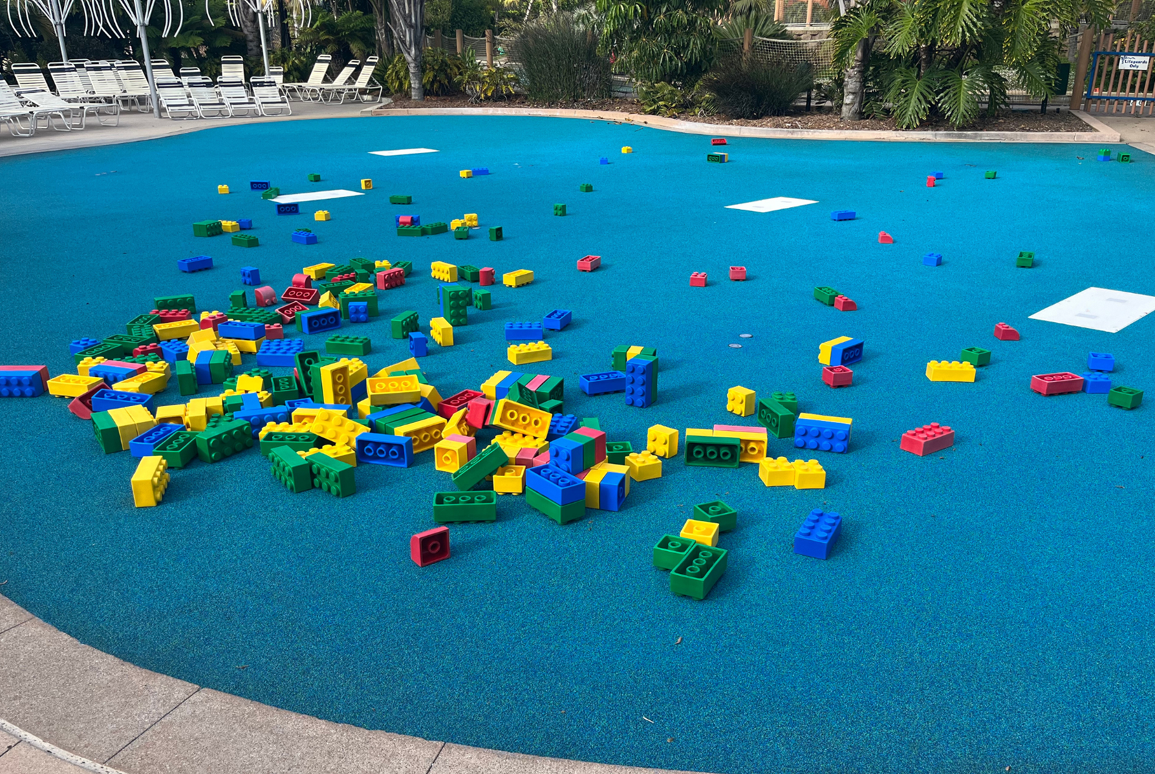 Colorful plastic building blocks scattered on a blue playground surface near poolside lounge chairs, surrounded by lush green plants and trees.