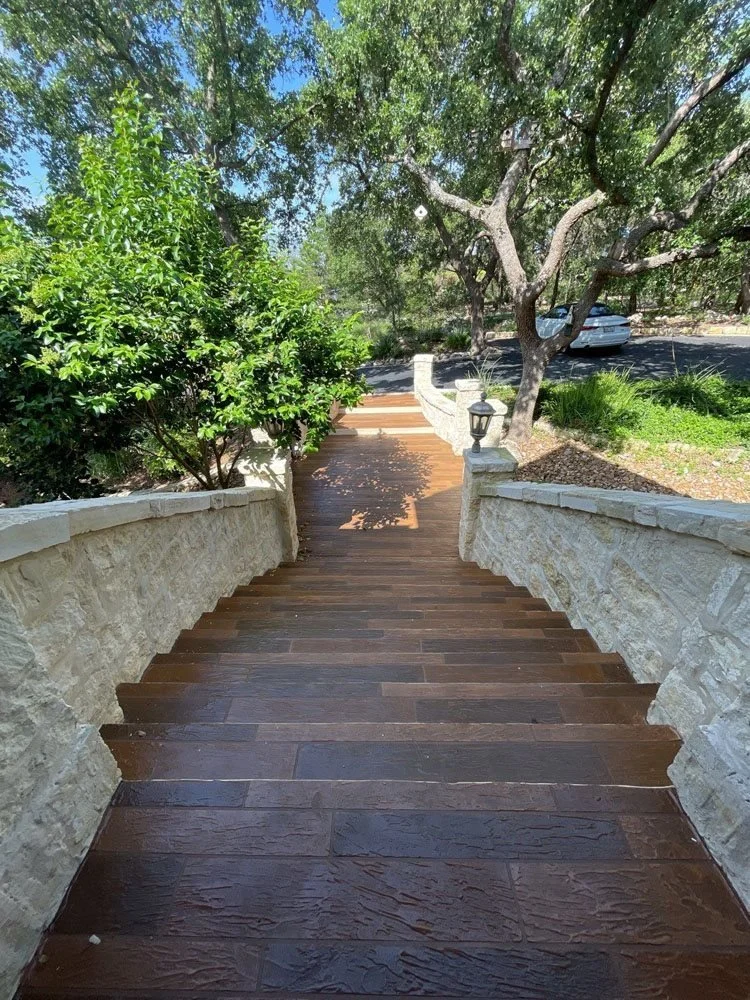 A wooden staircase with stone sidewalls leading down to a sidewalk, surrounded by green trees and shrubs in a sunny outdoor setting.