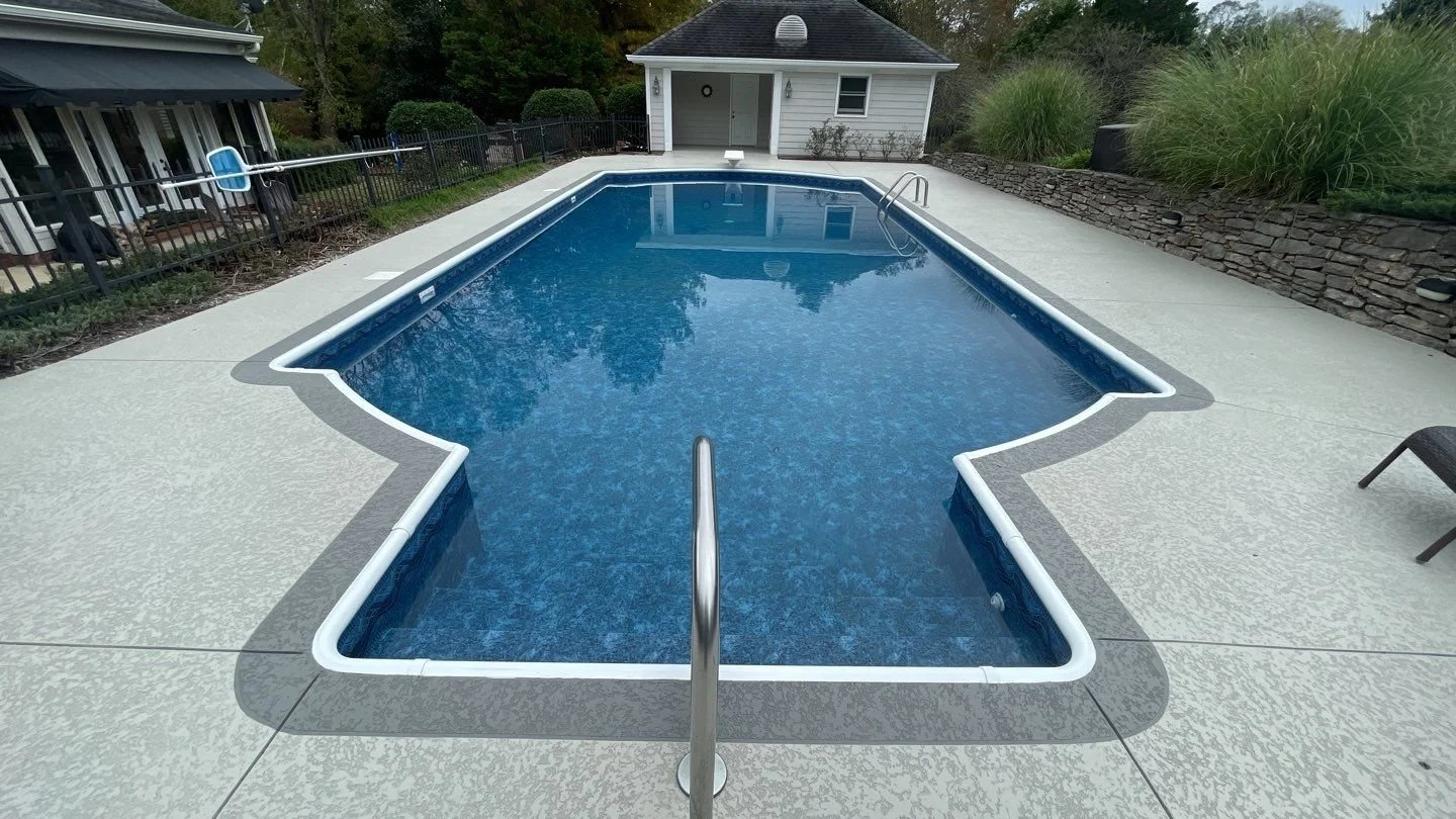 A rectangular backyard swimming pool with a blue interior, surrounded by a light-colored concrete deck and a black metal fence. There is a small pool house or shed at the far end, with lush greenery and shrubs around the area.