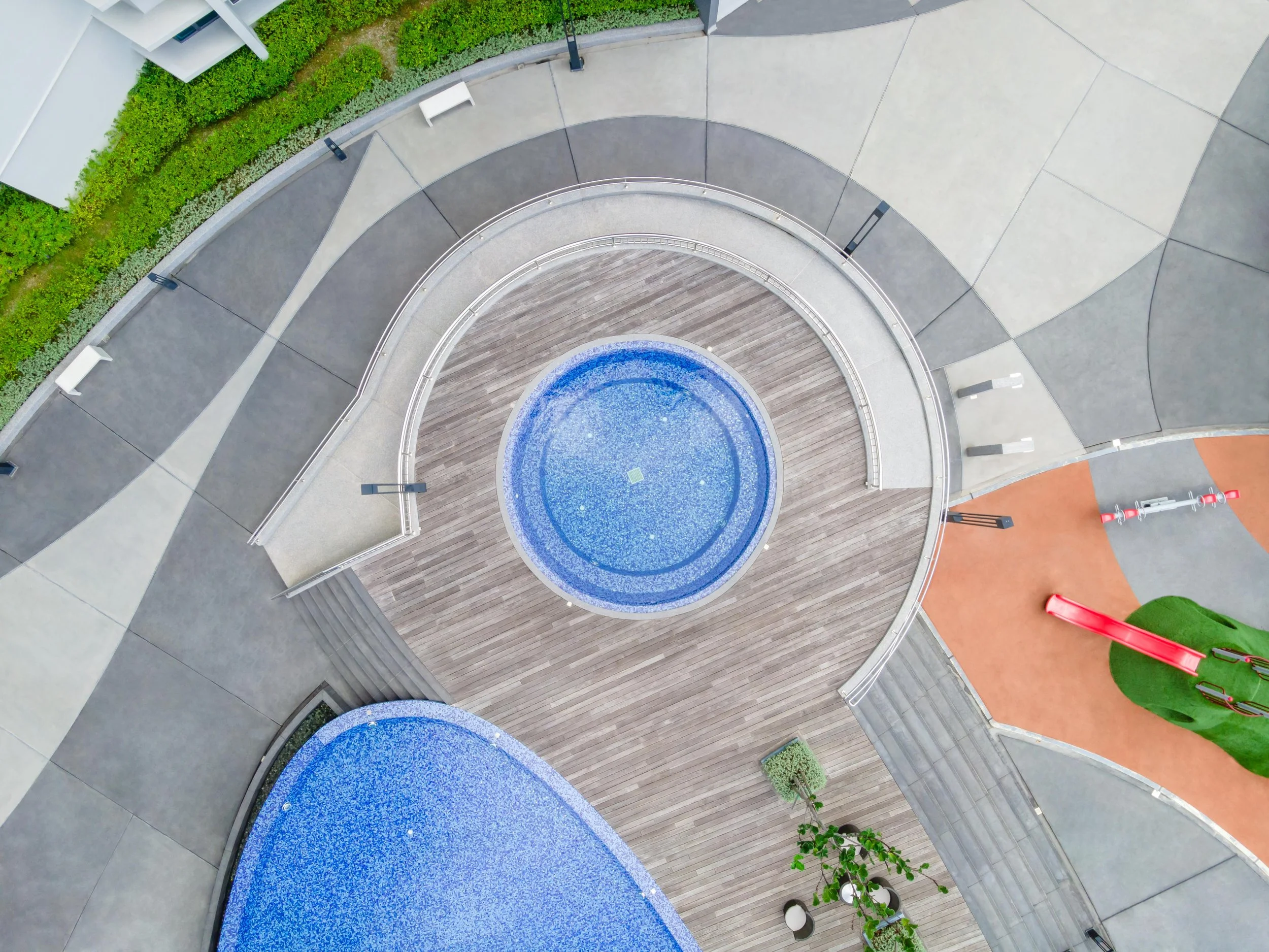 An overhead view of a courtyard with a small round blue fountain at the center, surrounded by wooden and concrete surfaces, benches, a green shrub, and an orange playground with a slide and tricycles.