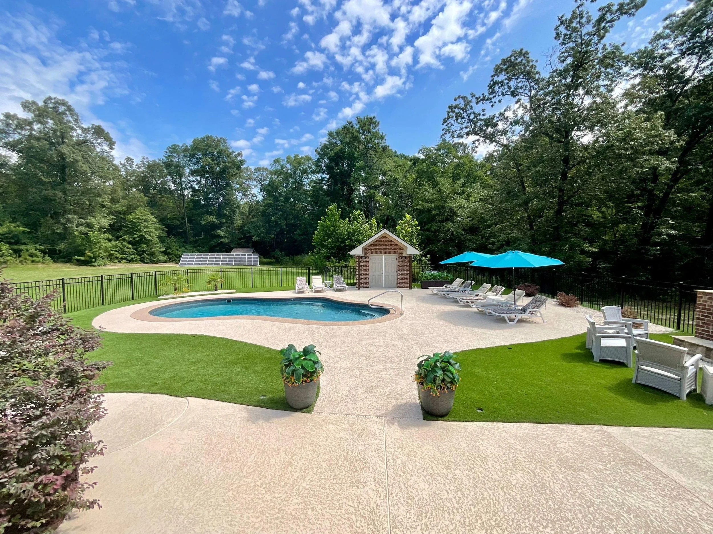 Residential backyard with a swimming pool, lounge chairs, large blue umbrella, and a small brick and white shed, surrounded by a black metal fence and lush green trees.