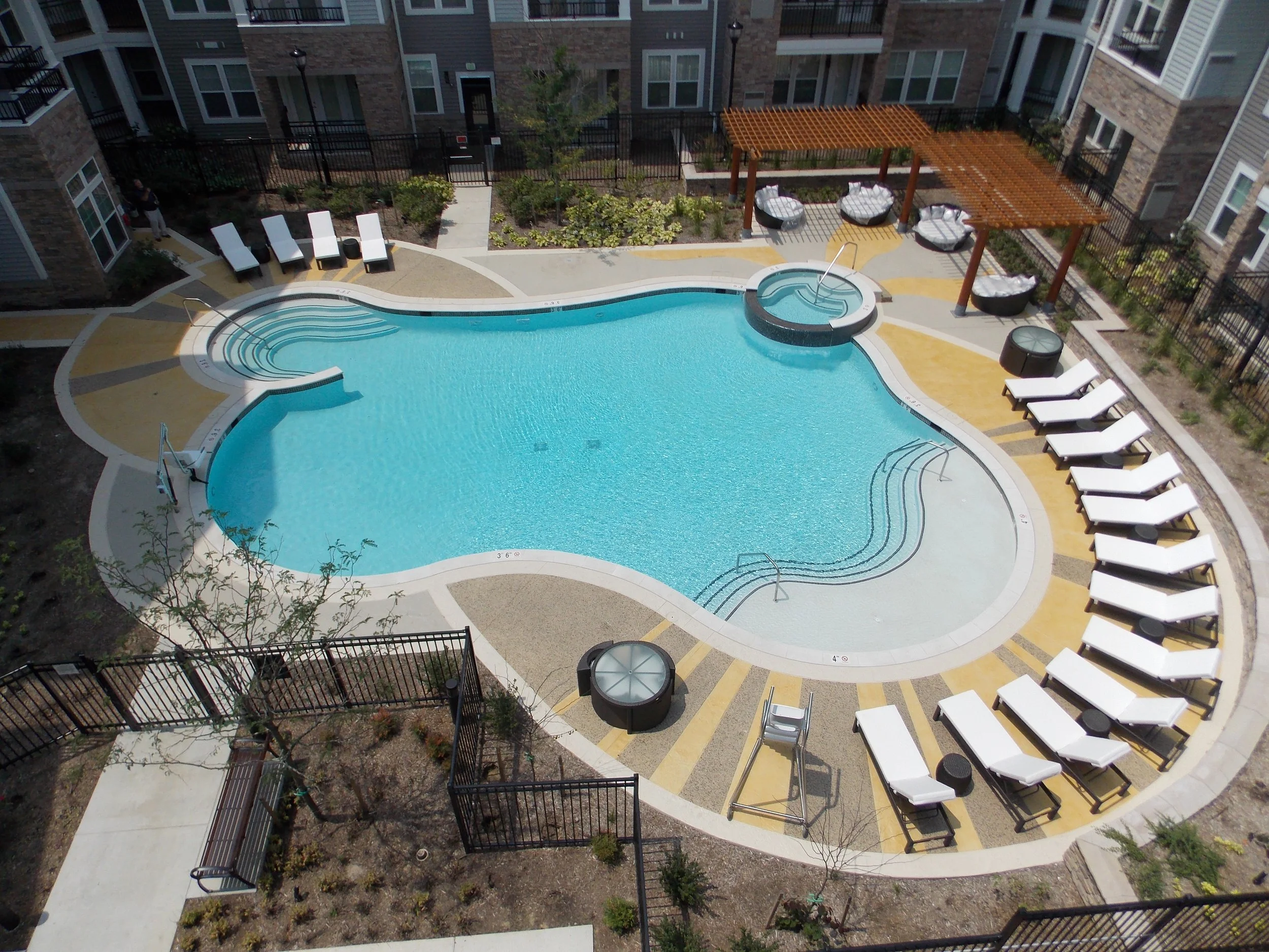 An aerial view of an outdoor swimming pool area in a residential complex, with lounge chairs, shaded cabanas, and a hot tub, surrounded by a fenced courtyard and apartment buildings.