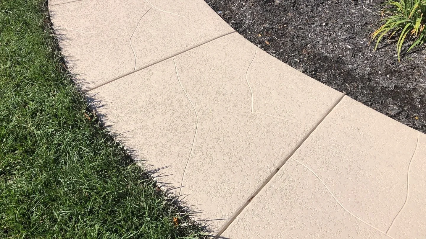 Close-up view of a sidewalk with beige textured tiles, surrounded by a grassy area on the left and a flower bed with dark mulch and green plants on the right.