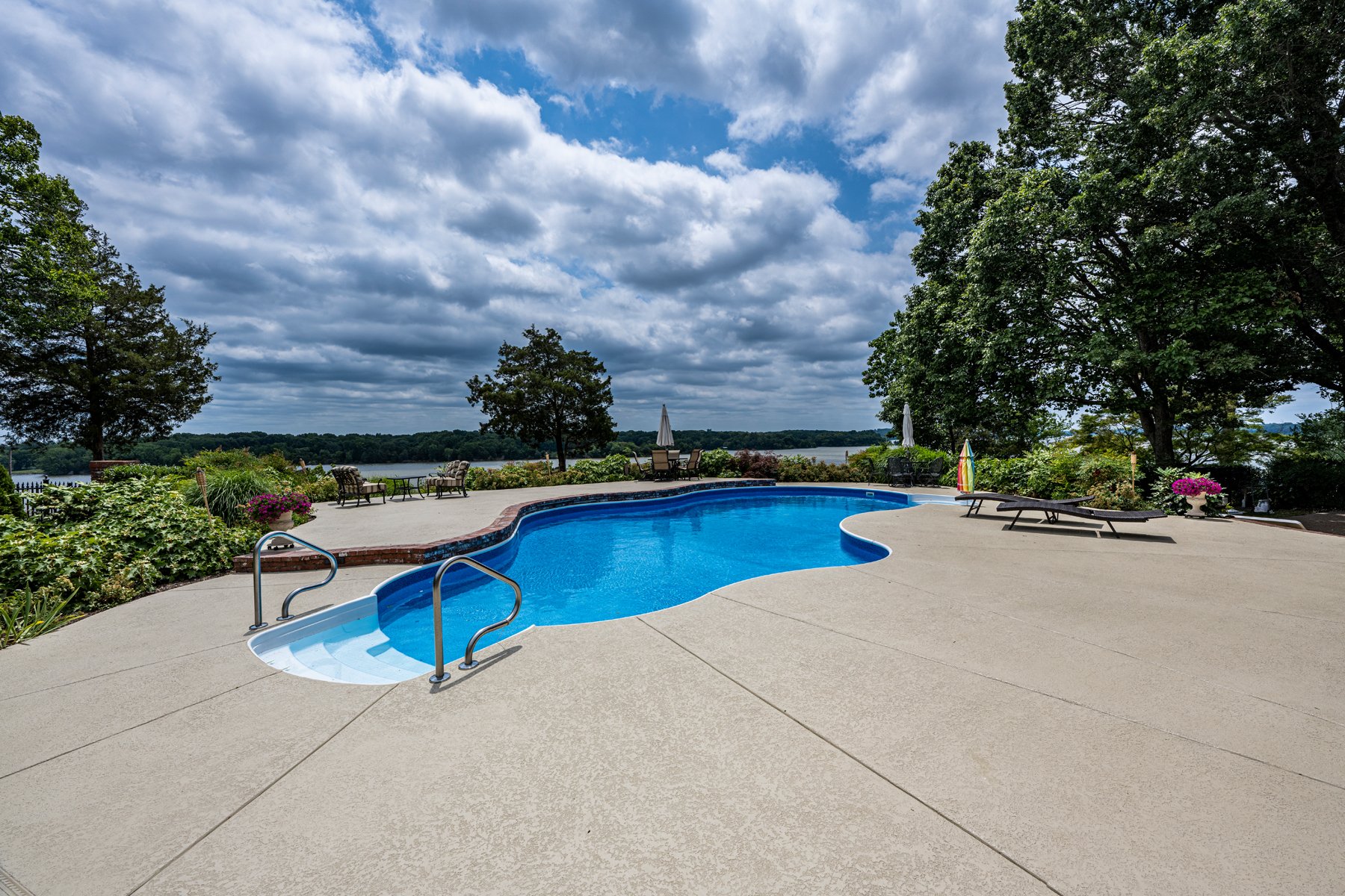 An outdoor swimming pool with a curved shape, surrounded by a concrete deck and green trees. There are lounge chairs, umbrellas, and benches around the pool, overlooking a body of water in the background under a partly cloudy sky.
