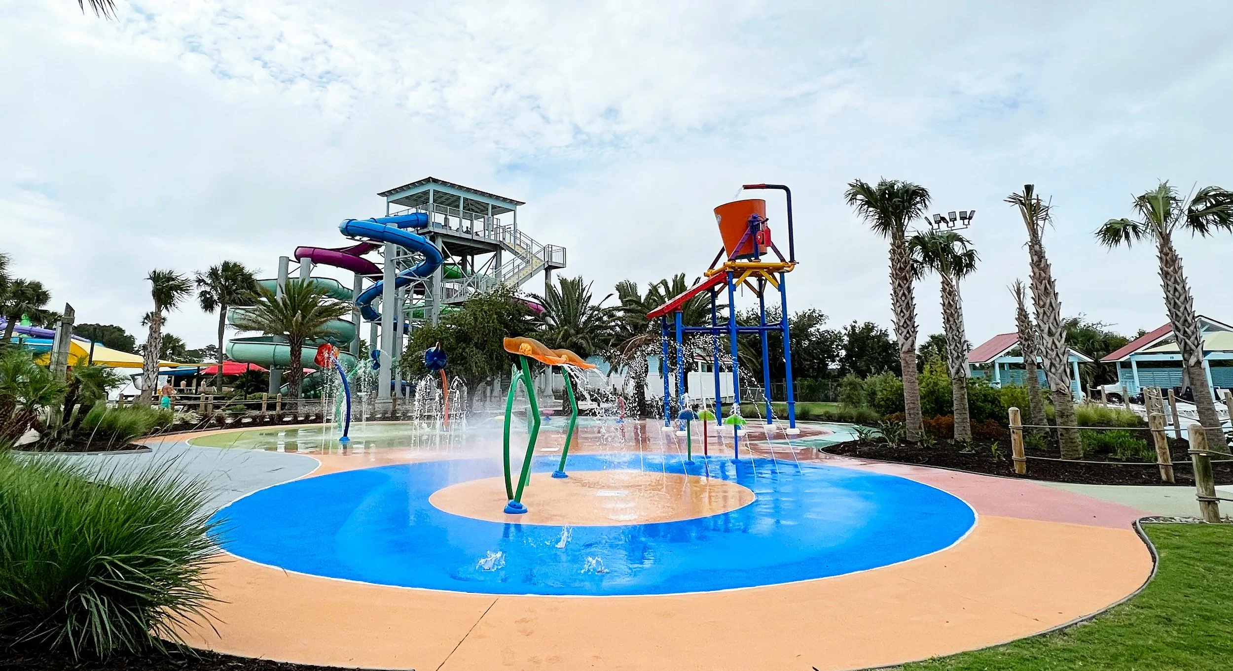 Colorful water play area with slides, fountains, and water sprinklers, surrounded by palm trees and a water slide structure in the background, under a partly cloudy sky.