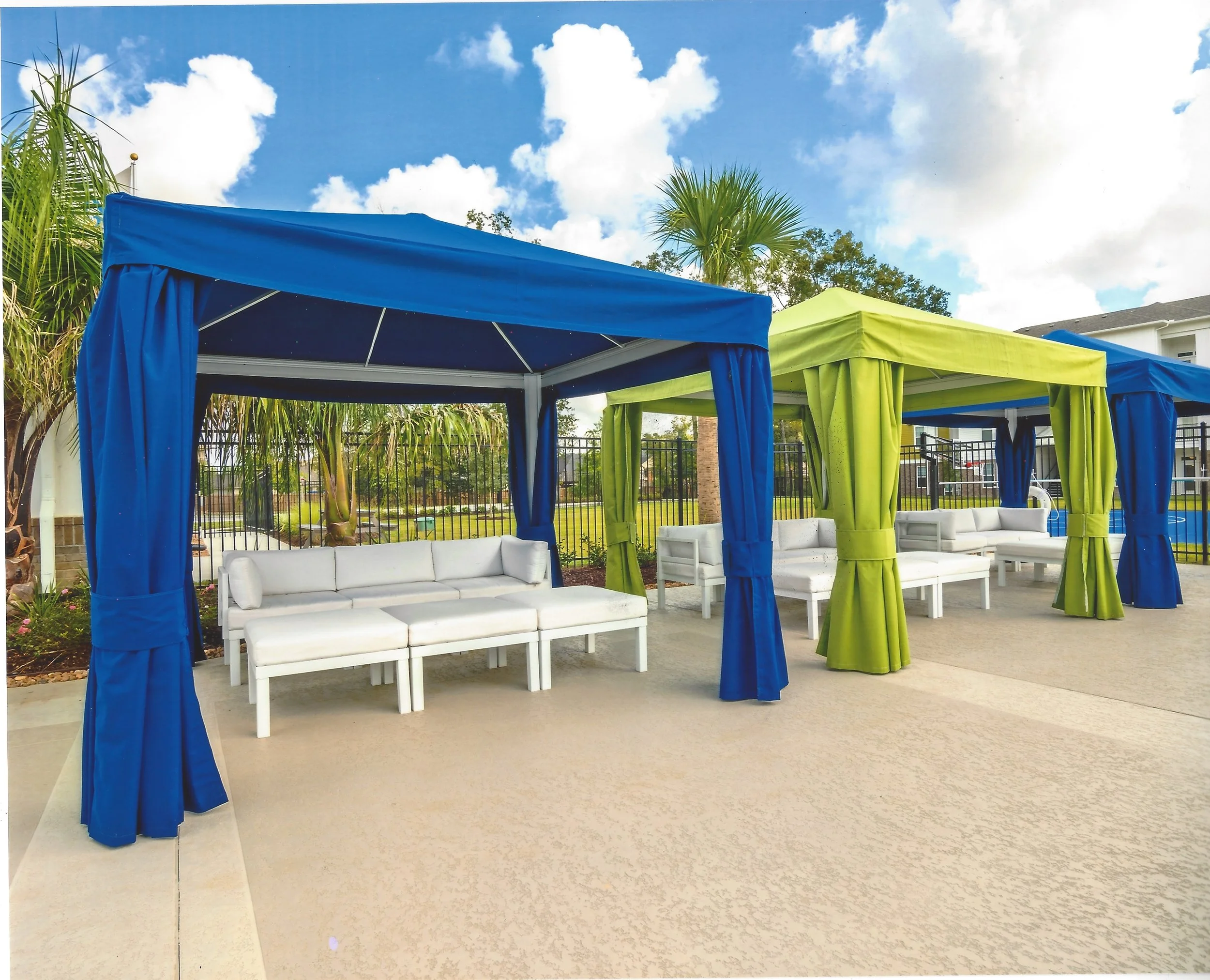 Colorful outdoor cabanas with blue and green curtains, white sofas, and a clear sky with scattered clouds.