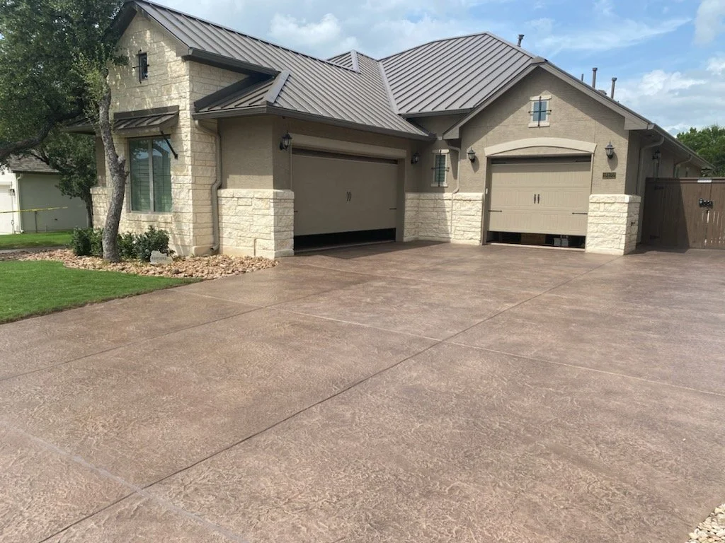 A residential house with a garage and a large driveway made of stamped concrete. The house has a stone facade and beige siding, with a metal roof. There are two garage doors, and small decorative windows above them. There are trees and landscaping wi