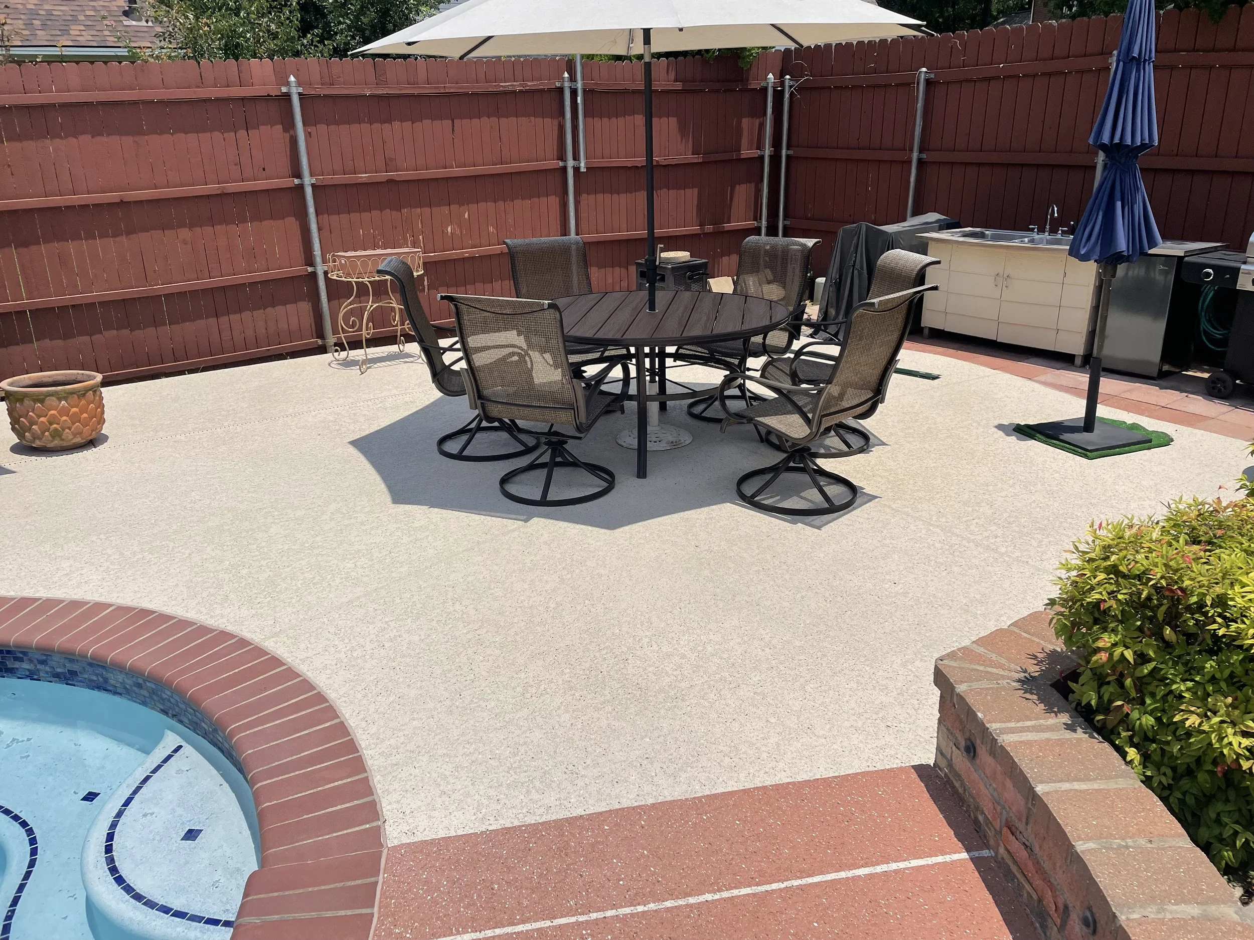 Backyard patio with a round table and six chairs, a large umbrella, a hot tub, a barbecue grill, and a flowerpot, enclosed by a red wooden fence.