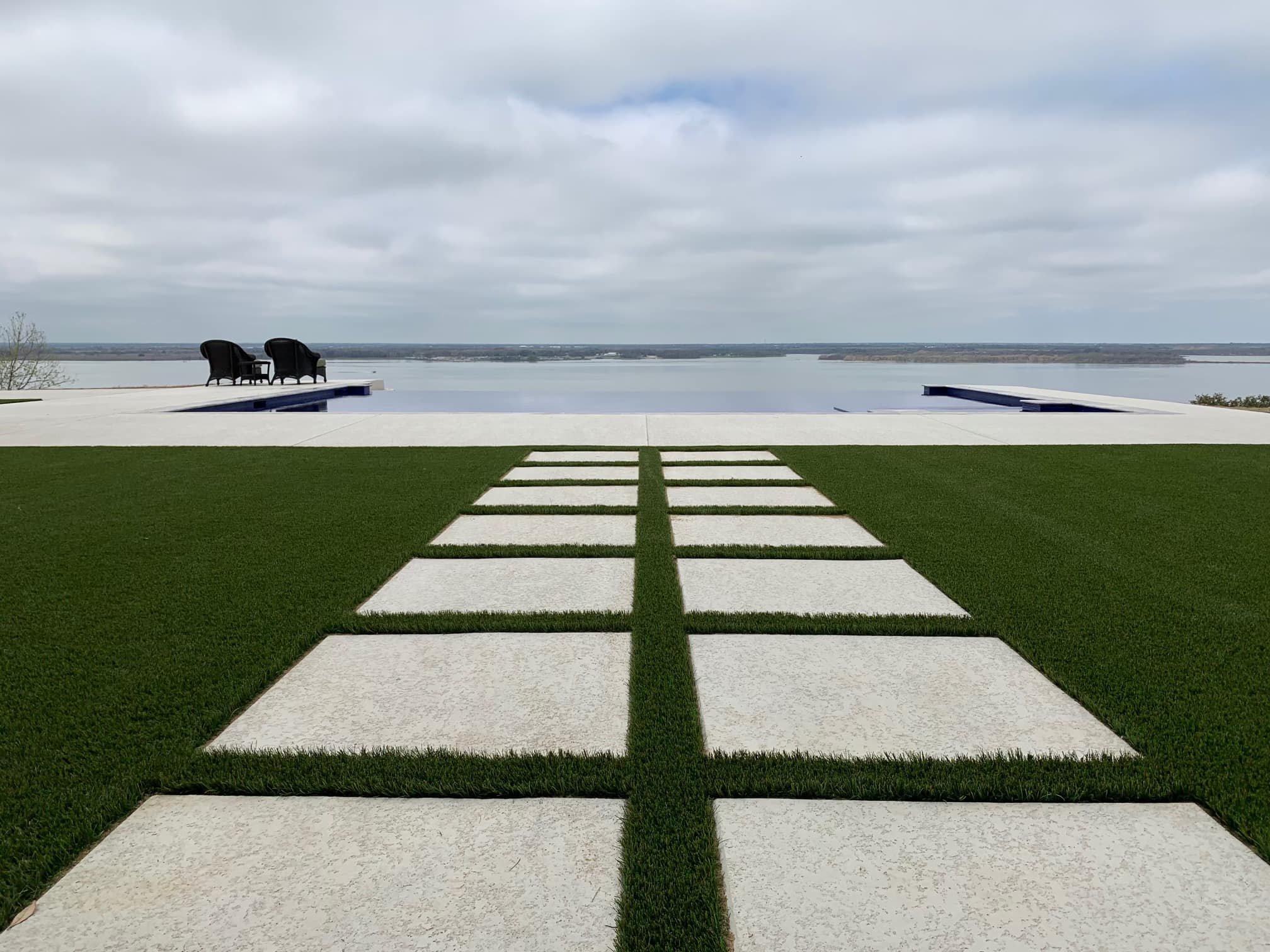 Modern backyard with a stone pathway leading to an infinity pool overlooking a lake. Two black chairs are positioned near the pool on a white patio. Overcast sky.
