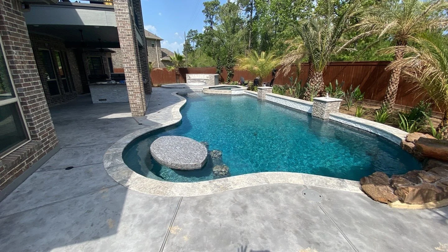 Backyard swimming pool with a curved shape, surrounded by concrete decking, with palm trees and plants along the fence in the background, and part of a house with brick exterior visible on the left.