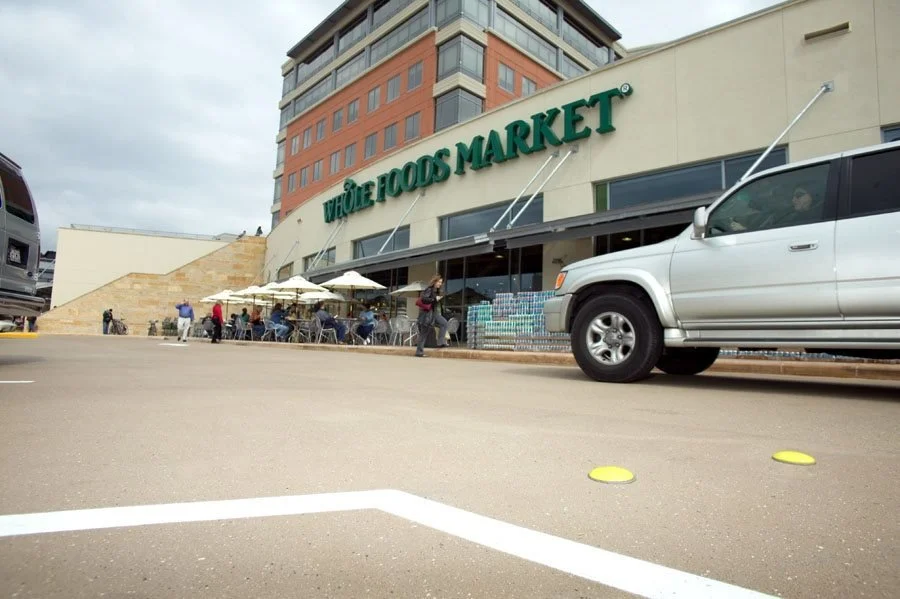 Exterior view of Whole Foods Market with outdoor seating, umbrellas, a white pickup truck parked nearby, shopping carts, and customers entering or exiting the store.