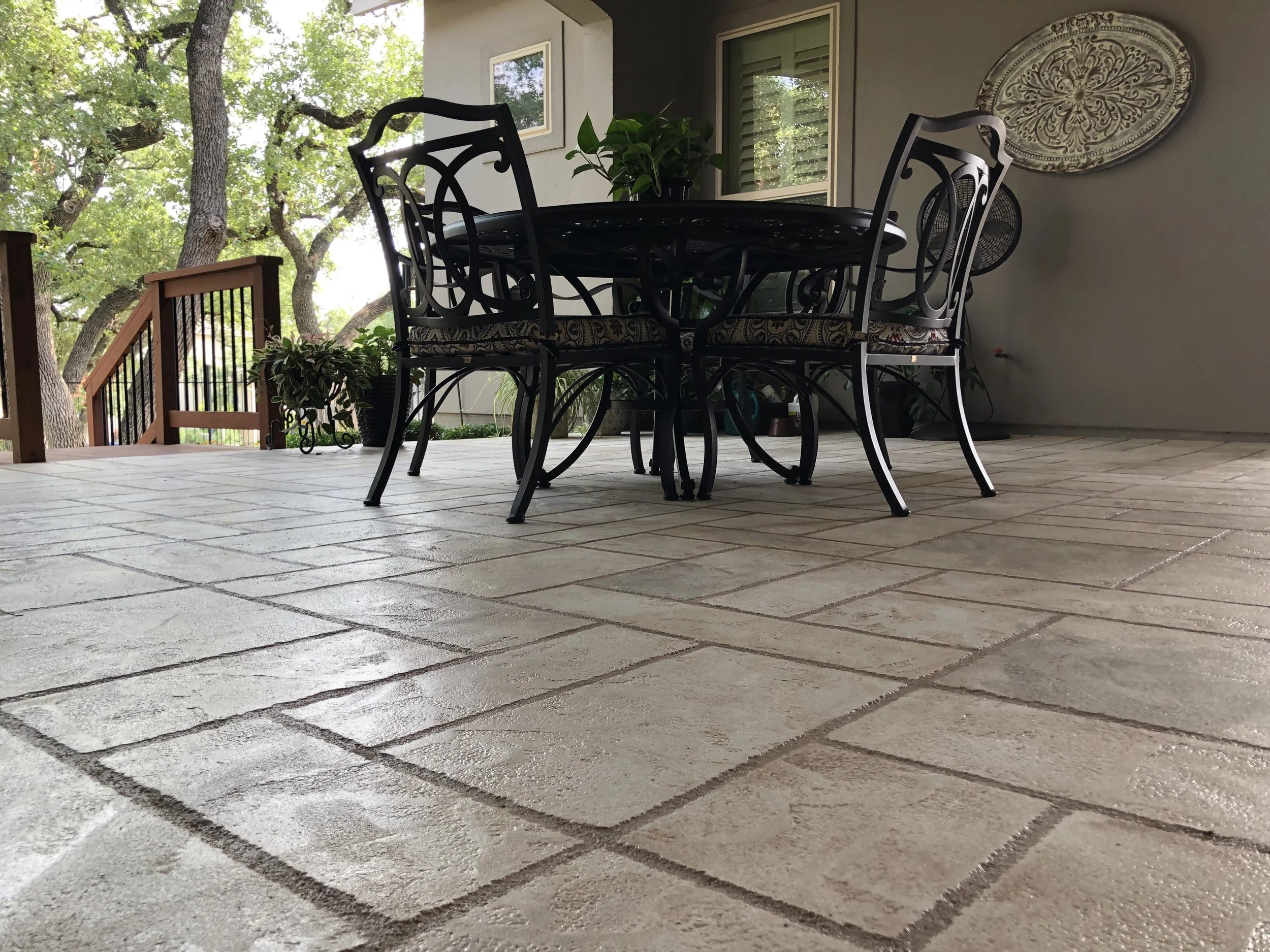 Outdoor patio featuring a black wrought iron table and chairs with a patterned cushion, surrounded by potted plants, trees in the background, and a beige tiled floor.