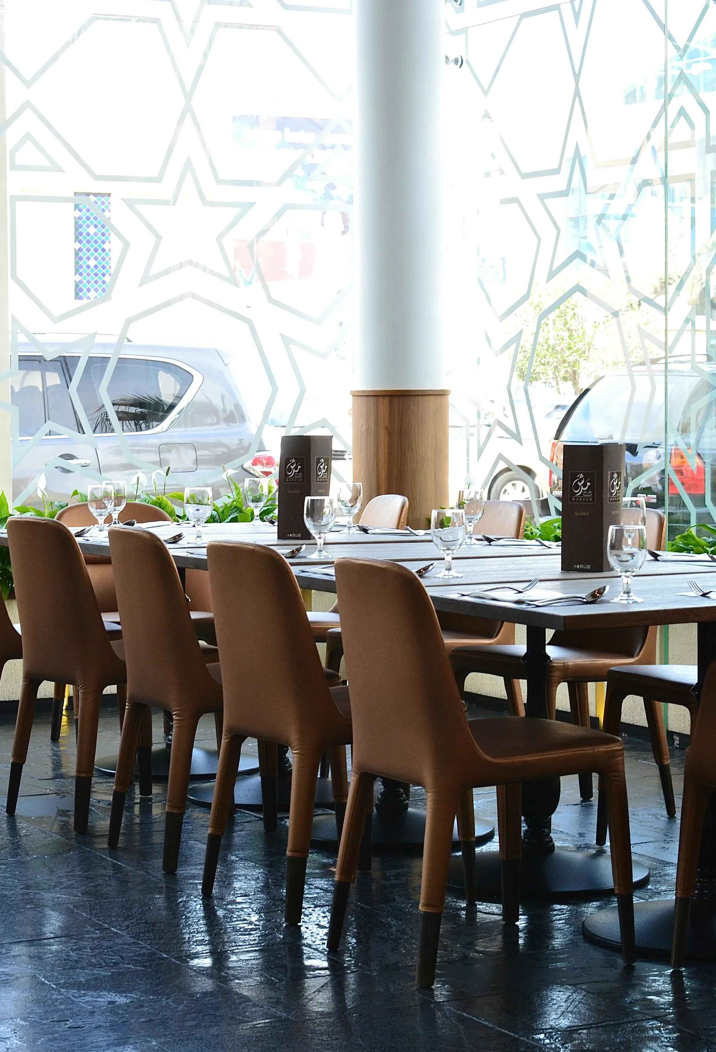 Empty restaurant with a long wooden table set with glassware, silverware, and black menus, surrounded by brown chairs, large windows with star-shaped decals, and parked cars outside.