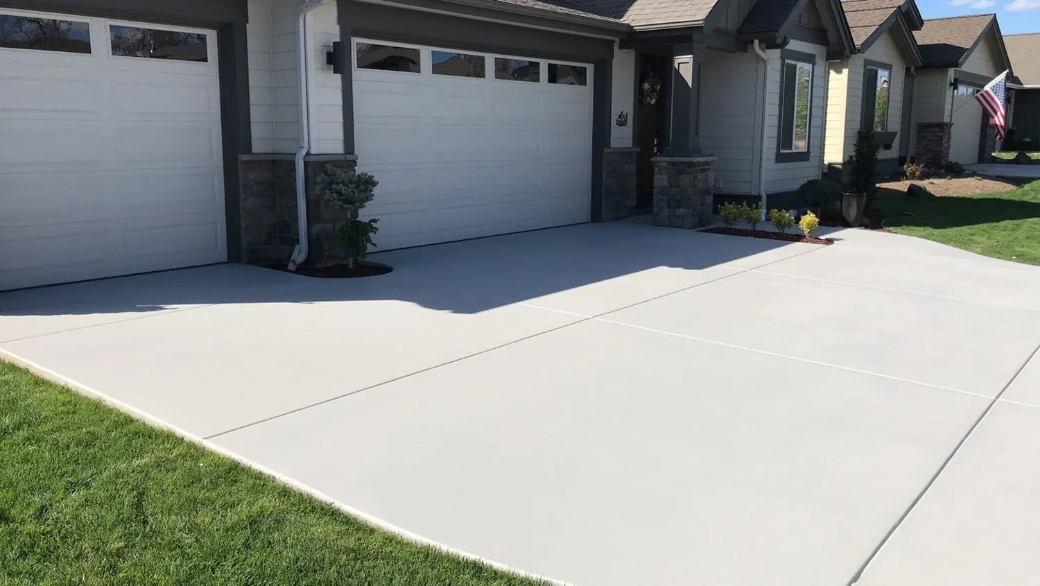 Clean concrete driveway in front of a modern house with a two-car garage and a small garden with plants and American flag in the background.