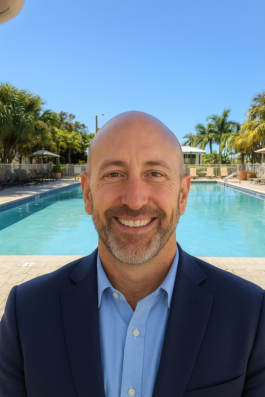 A smiling man in a blue suit and light blue shirt standing in front of a swimming pool with palm trees and umbrellas under a clear blue sky.