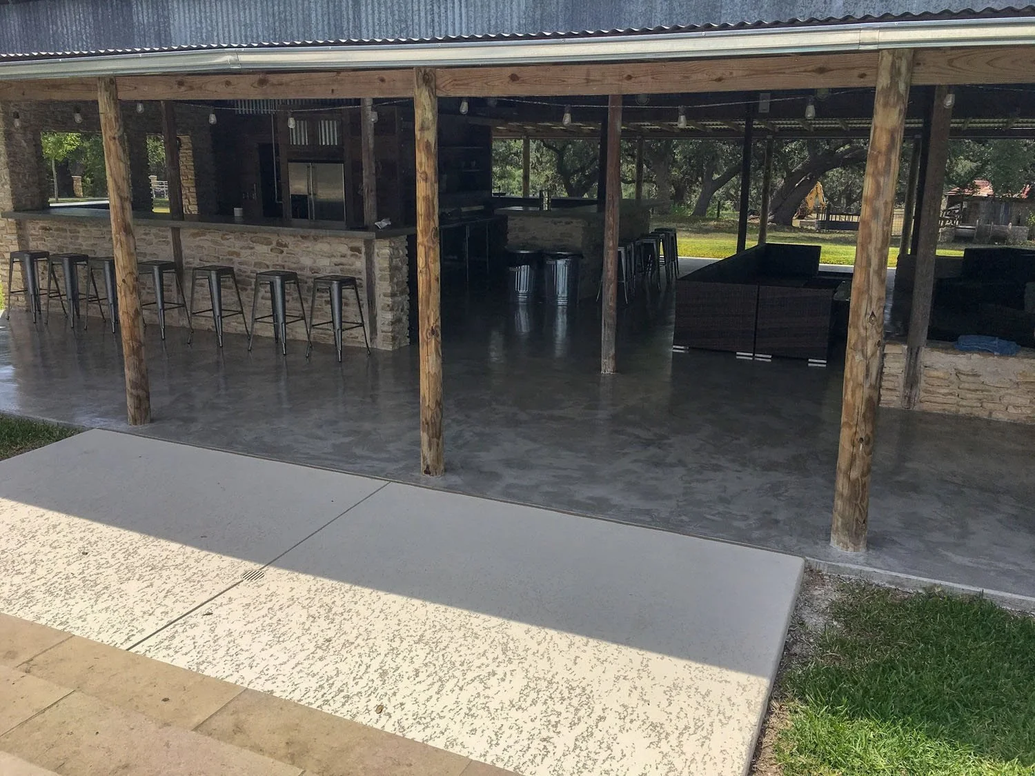 Outdoor covered bar area with wooden posts, a stone counter with bar stools, and a sitting area with dark furniture, overlooking a grassy yard and trees.