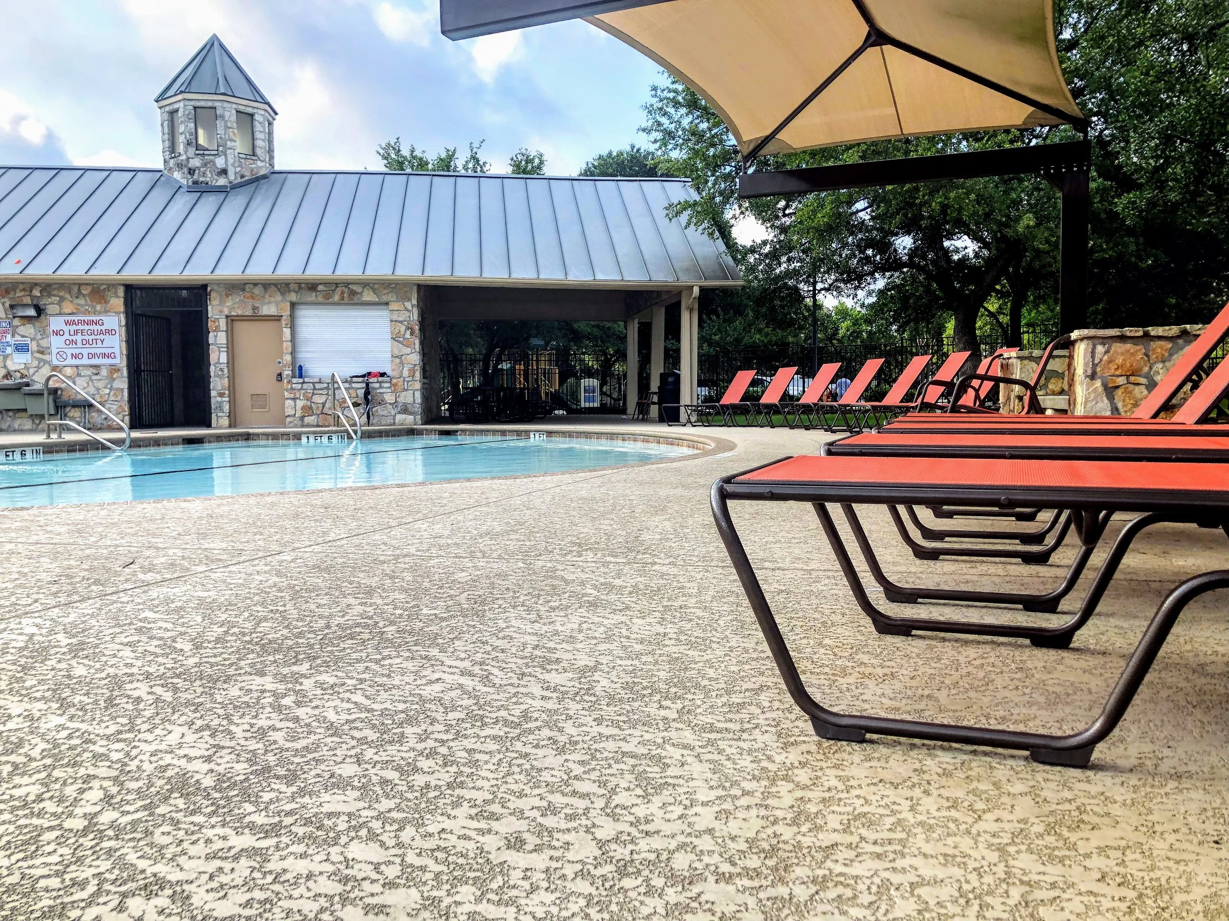 An outdoor swimming pool area with red lounge chairs, a large umbrella, and a small stone building with a metal roof in the background. Trees surround the area.