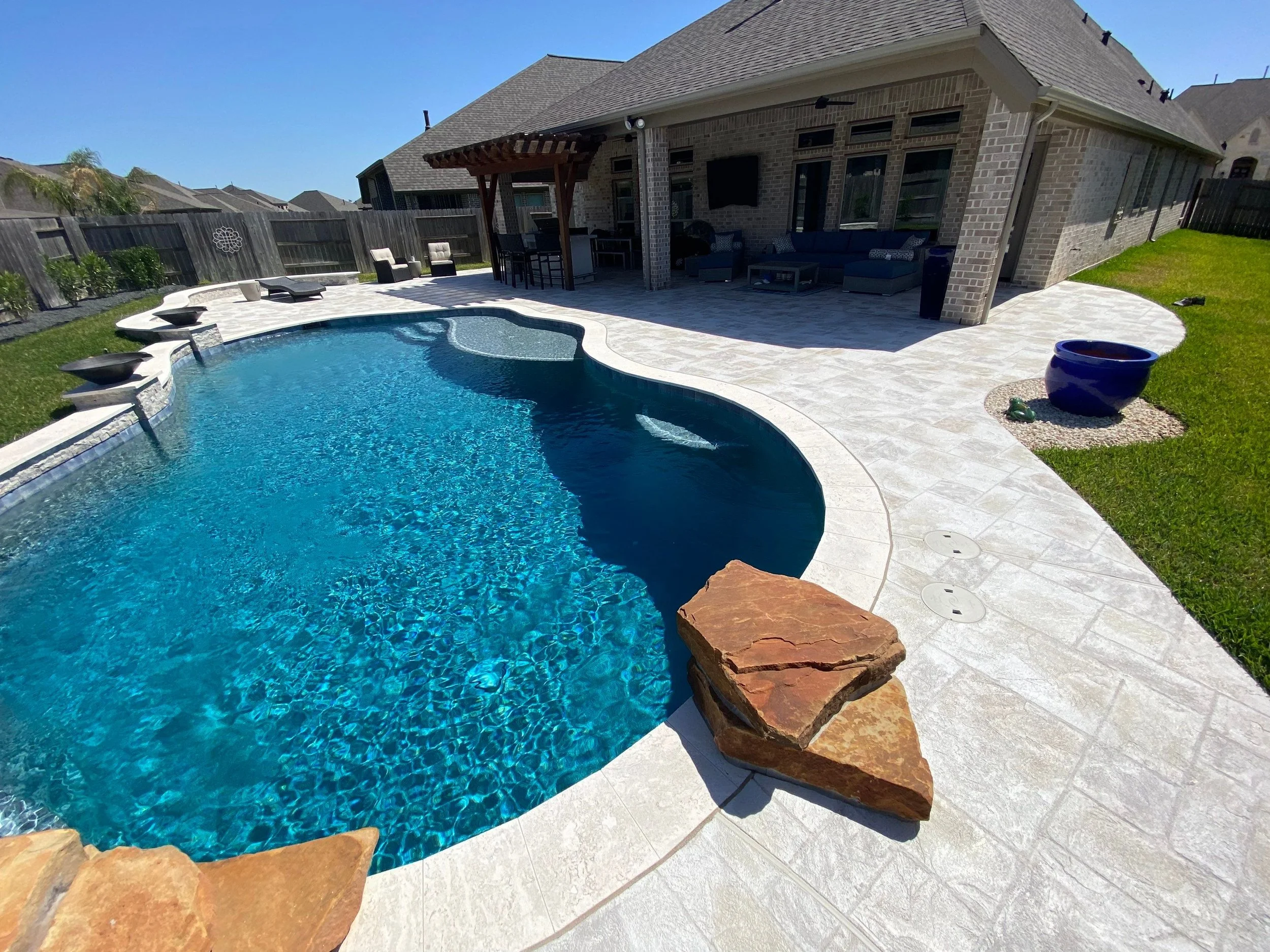 Backyard with in-ground swimming pool, poolside seating, a covered patio area with outdoor furniture, and a brick house in the background.