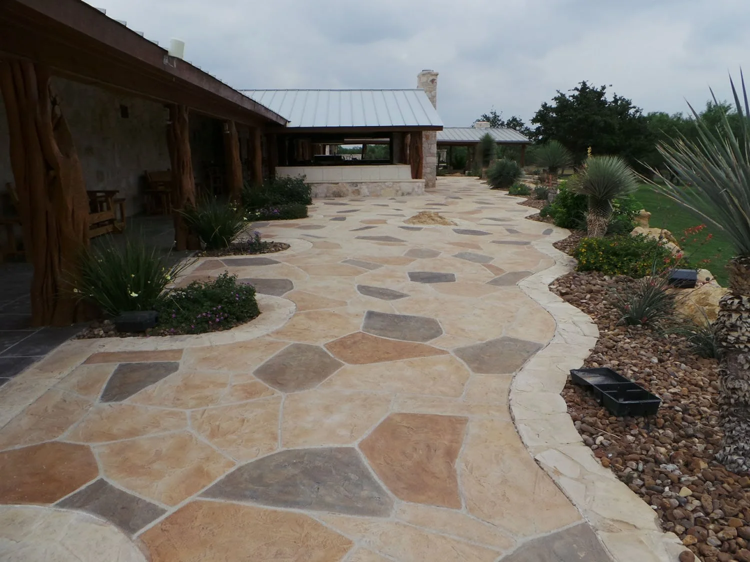 A stone patio with tan, beige, and gray irregular stones, bordered by desert plants and small trees, with a covered wooden structure with seating on one side and a cloudy sky above.
