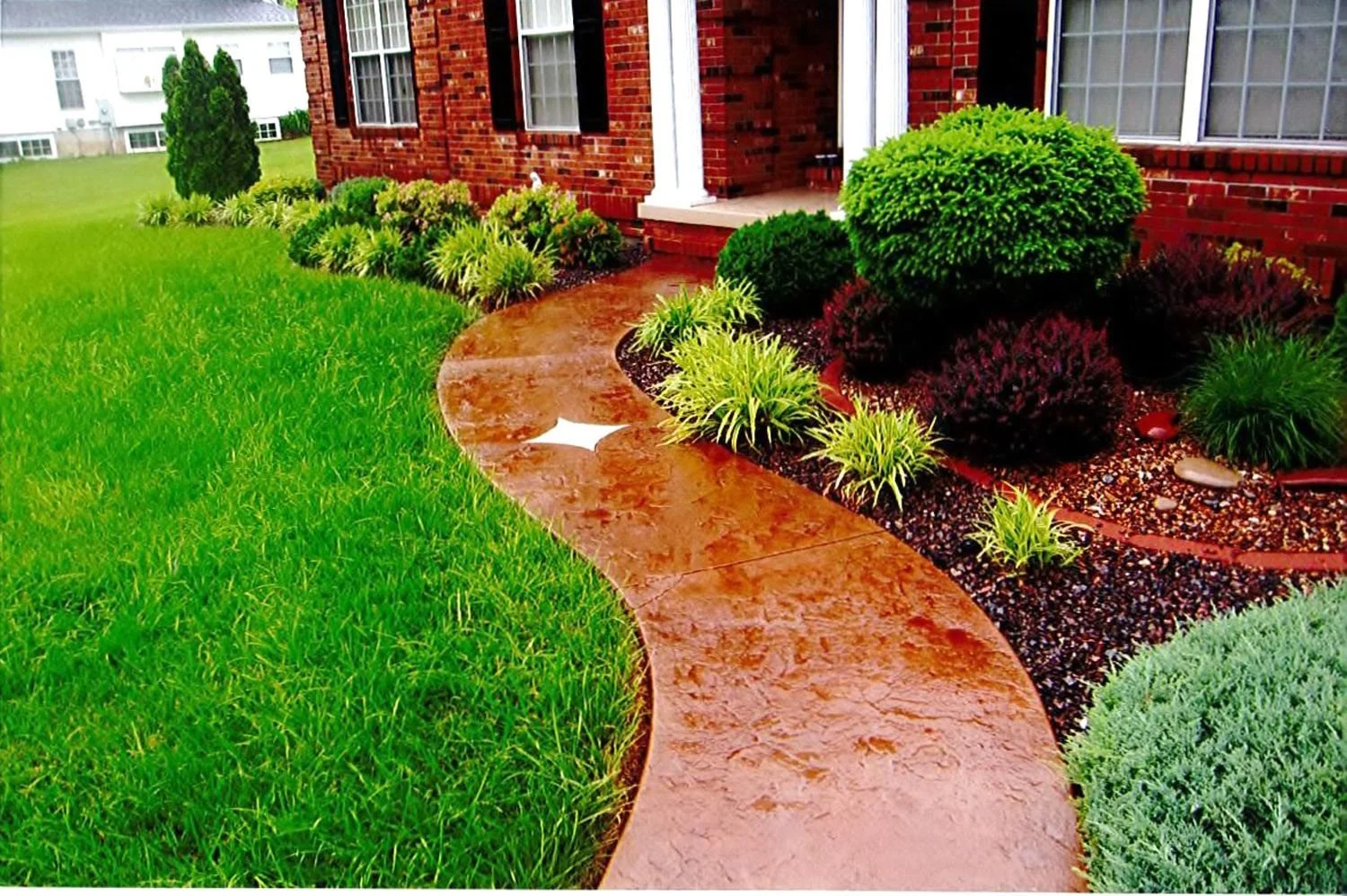Curved red concrete sidewalk leading to front door of a red brick house, surrounded by well-manicured green lawn and lush garden with various bushes and plants.