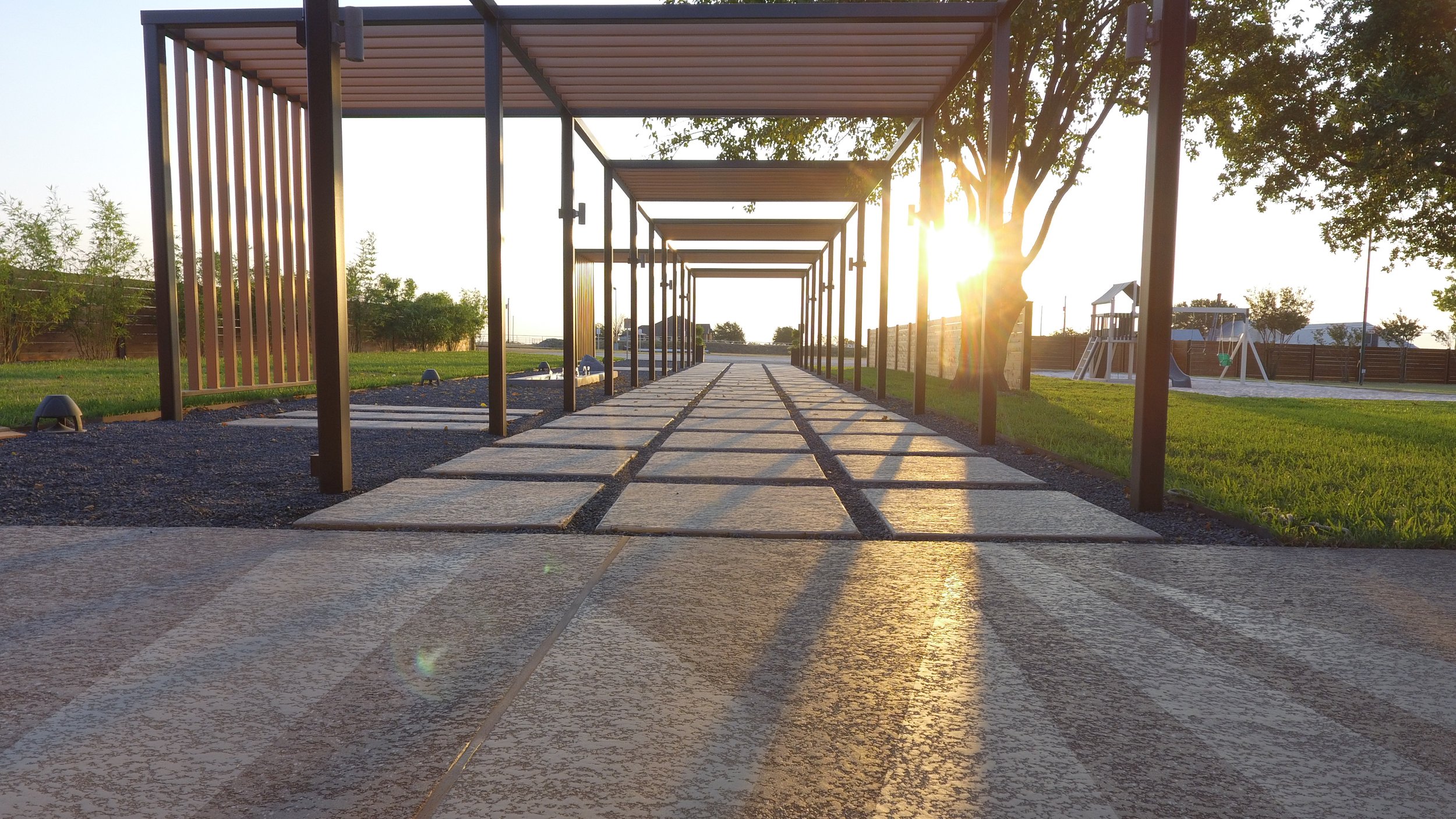 Pathway with rectangular stone tiles leading toward a sunset, with a wooden pergola overhead, on a grassy yard with trees, a swing set, and a fence